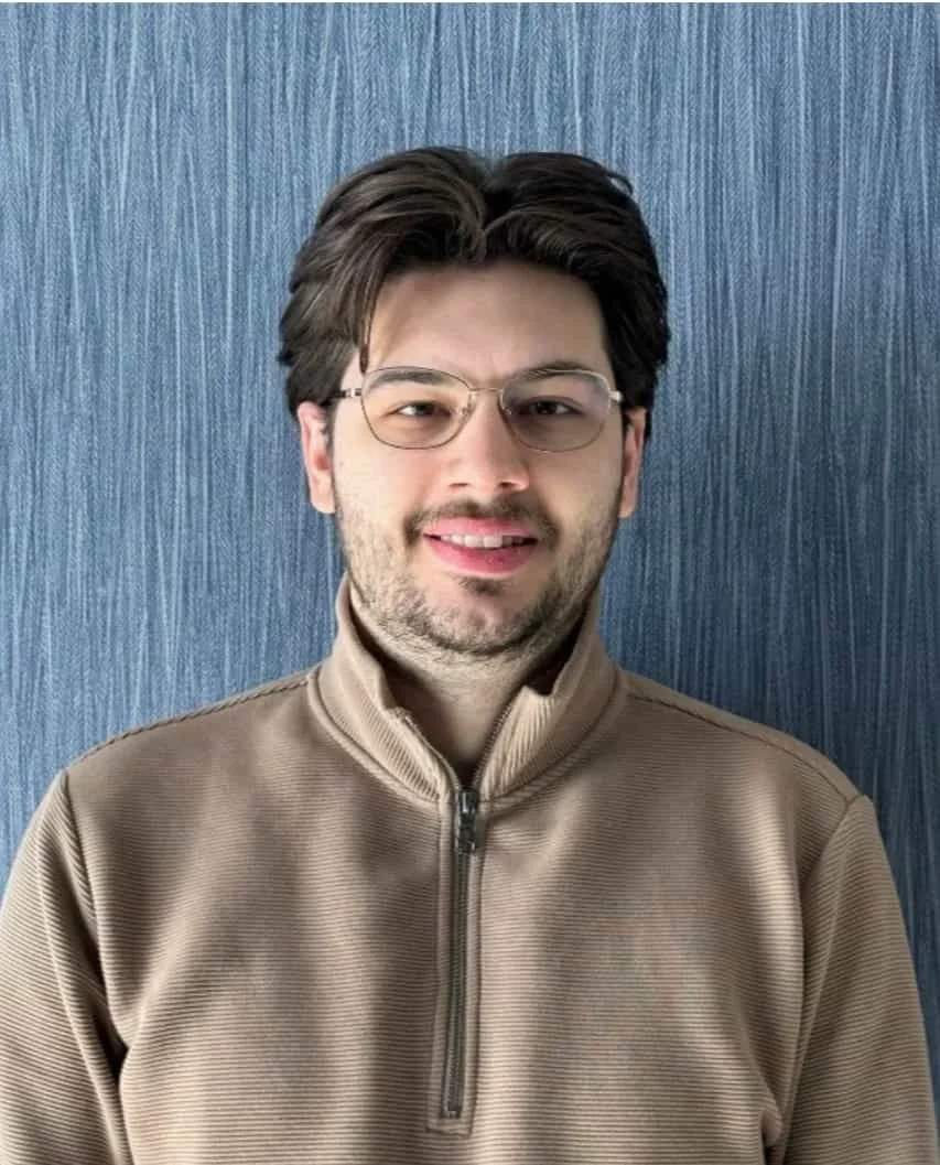 A man with glasses and brown hair, wearing a tan zip-up jacket, standing in front of a textured blue wall.
