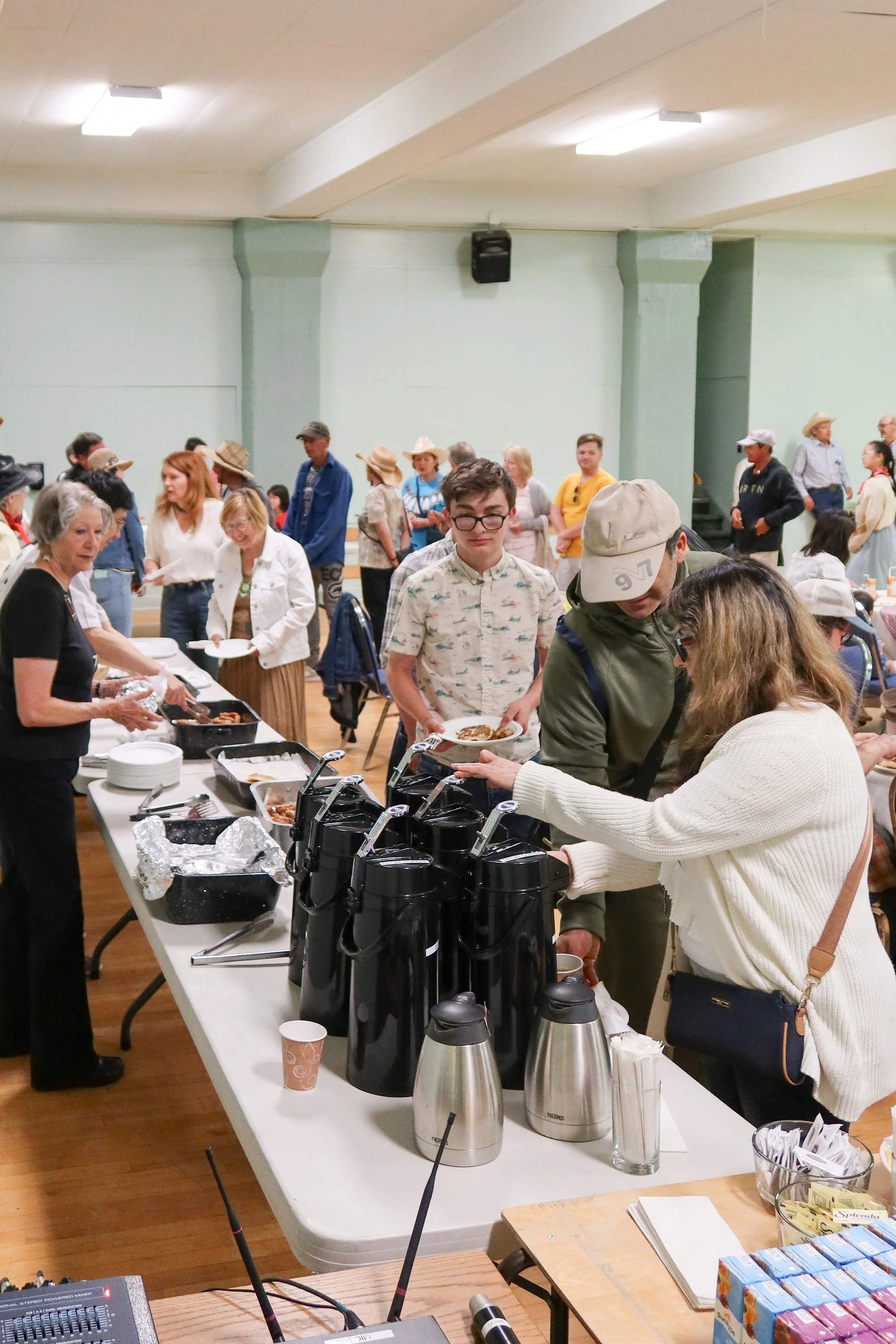 People lining up at a buffet with food and drink dispensers in a gymnasium.