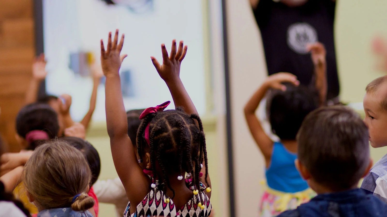 Children in a classroom raising their hands during a lesson, with a teacher at the front and a whiteboard