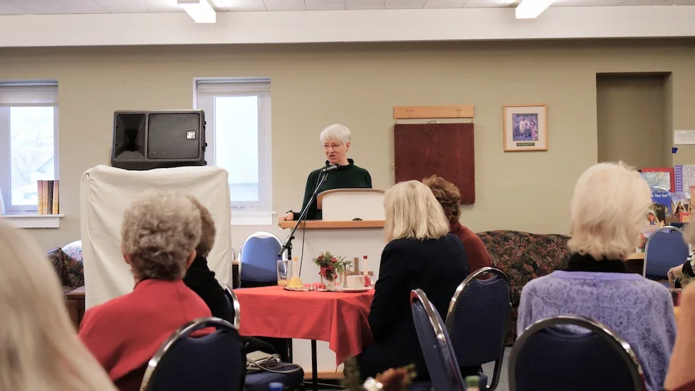 A woman standing at a podium speaking into a microphone in a room with an audience of mostly older women, some with grey hair, seated around tables with red tablecloths and small floral centerpieces.