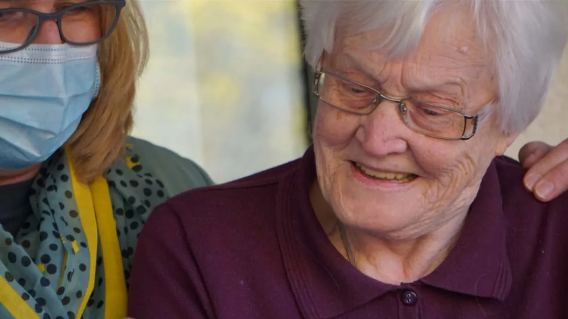 An elderly woman smiling and wearing glasses, with a caregiver beside her. The caregiver is wearing glasses, a face mask, and a polka dot shirt. The elderly woman has white hair and is wearing a maroon top.