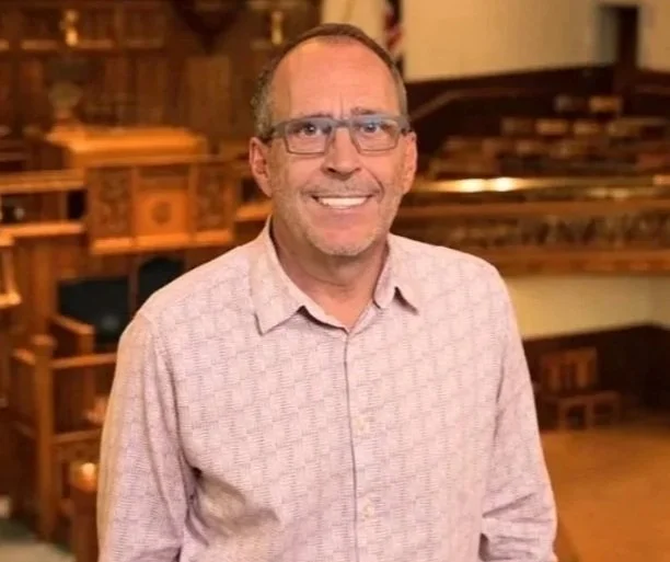 Man with glasses and a light pink checkered shirt smiling indoors, with wooden shelves and a piano in the background.