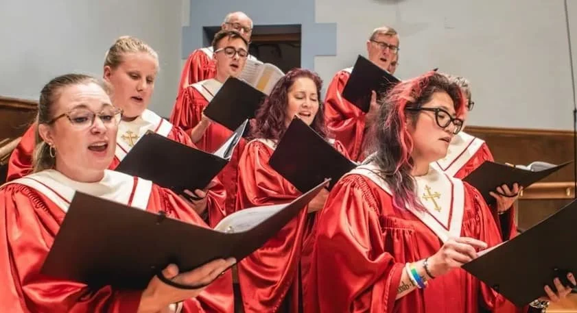 A choir of people in red robes singing and holding black folders in a church or concert hall.
