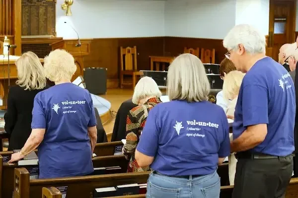 Group of volunteers in blue t-shirts with 'volunteer' printed on the back, standing and praying in a church pews.