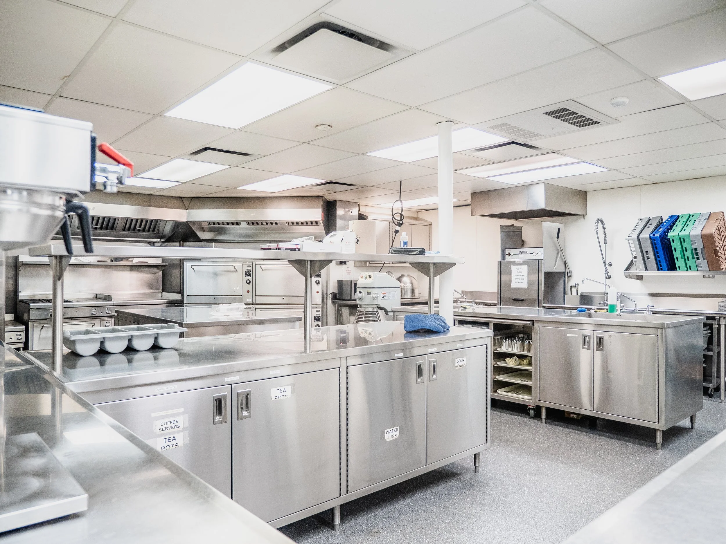 Empty commercial kitchen with stainless steel appliances, counters, and cabinets, including a coffee station, dishwasher, and shelves with various kitchen tools.