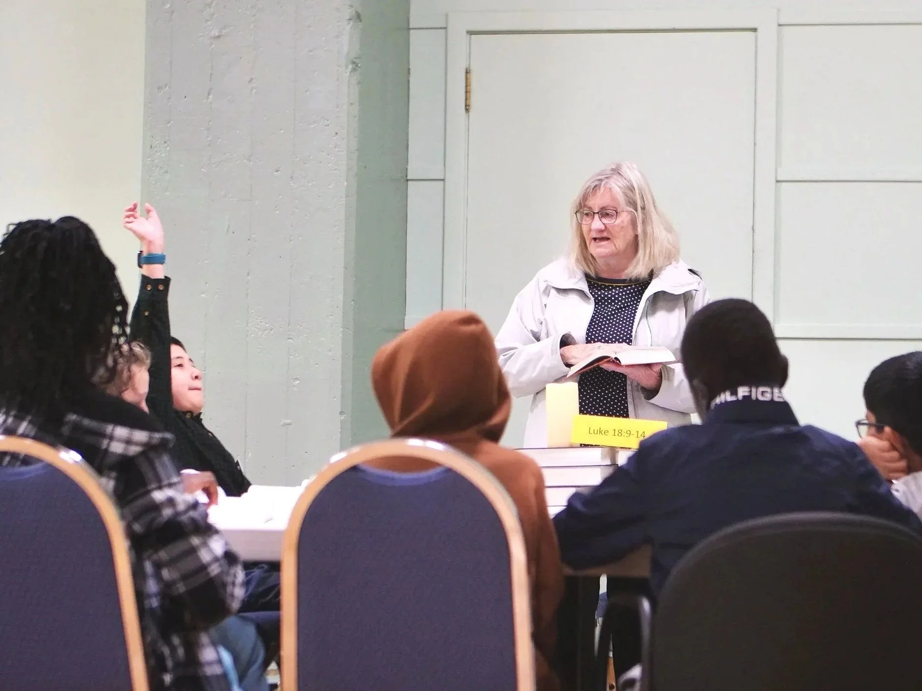 An woman teaching a Bible lesson to a diverse group of children and teenagers in a classroom or church setting, with a Bible open in her hands.