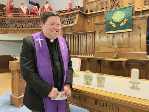 A clergyman in a black suit and purple stole standing in front of a church altar with candles and pottery, inside a church with wooden architecture and a stained glass window.