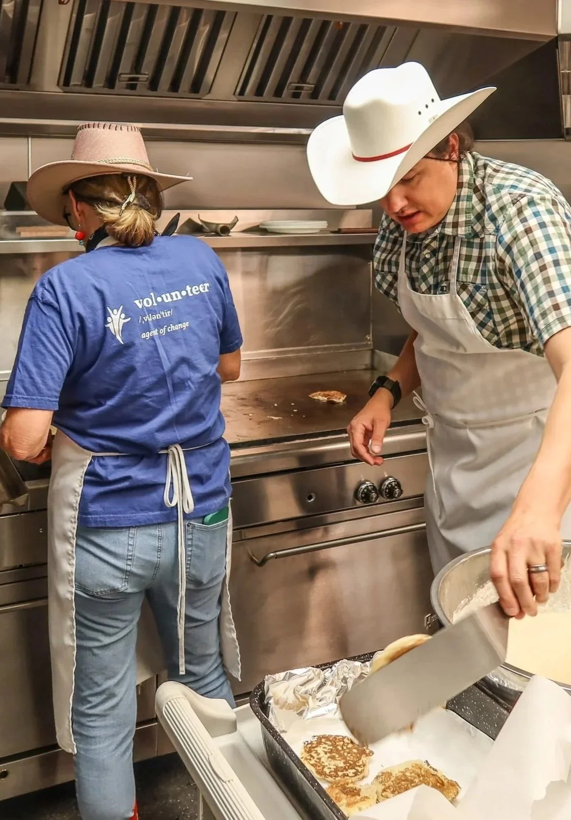 Two people cooking on a commercial stove, with pancakes in a tray in front of them, one wearing a cowboy hat and the other wearing a wide-brimmed hat, in a professional kitchen.