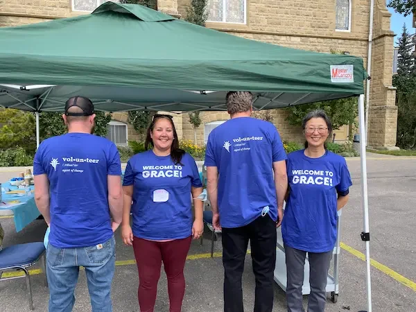 Four volunteers standing under a green canopy tent, wearing blue T-shirts that say "vol-un-teer" and "Welcome to Grace!" at an outdoor event in front of a building.