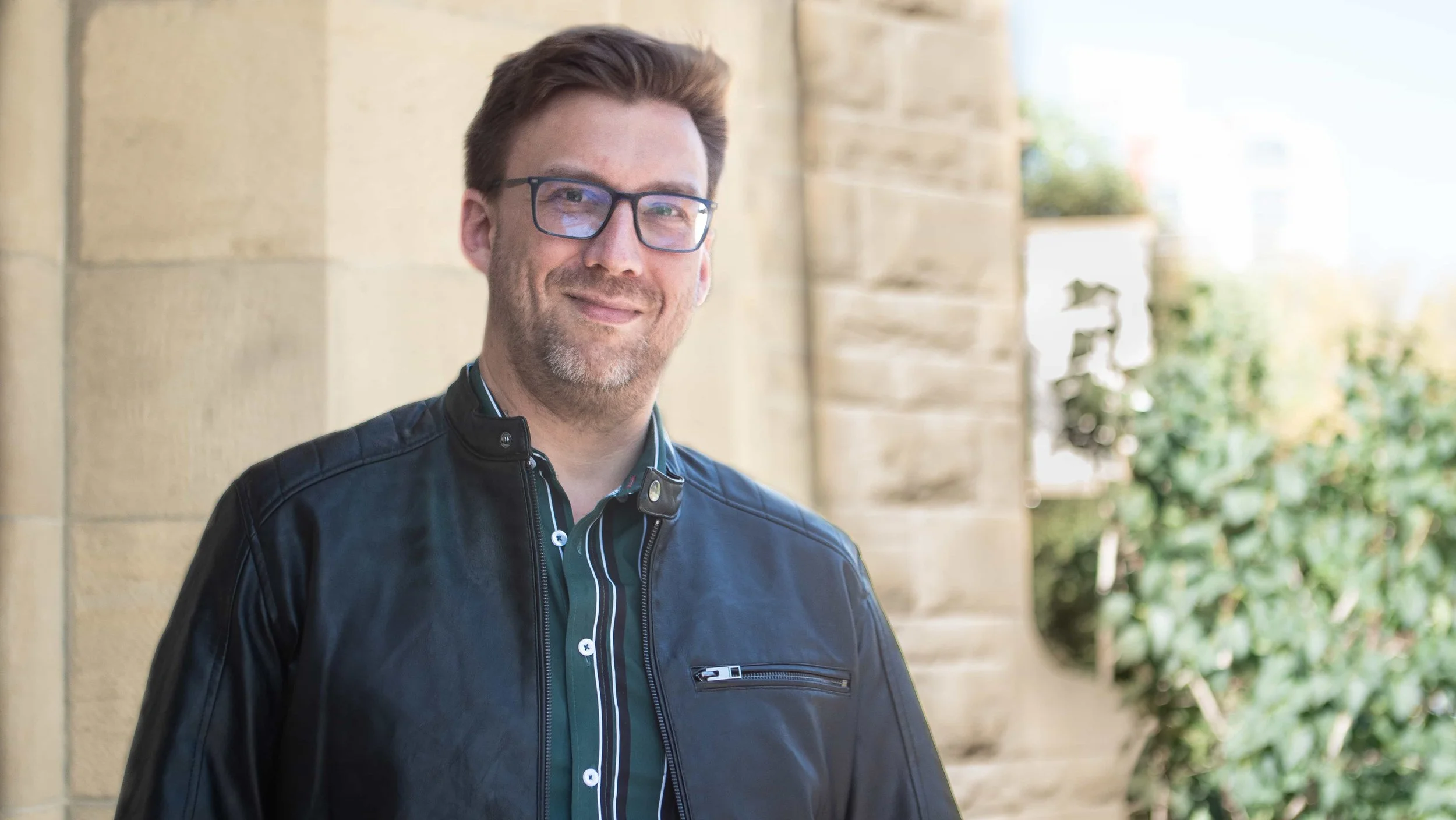 A smiling man with glasses, wearing a black leather jacket and a striped shirt, standing outdoors near a stone building and some greenery.