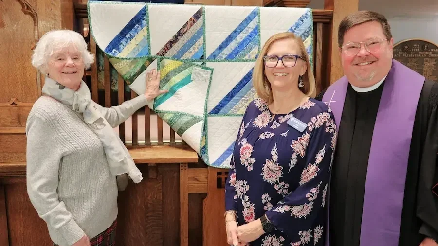 Three people standing in front of a quilt display, smiling. The woman on the left touches the quilt, the woman in the middle and man on the right are posing for the camera. The woman on the right wears a clerical collar and purple stole.