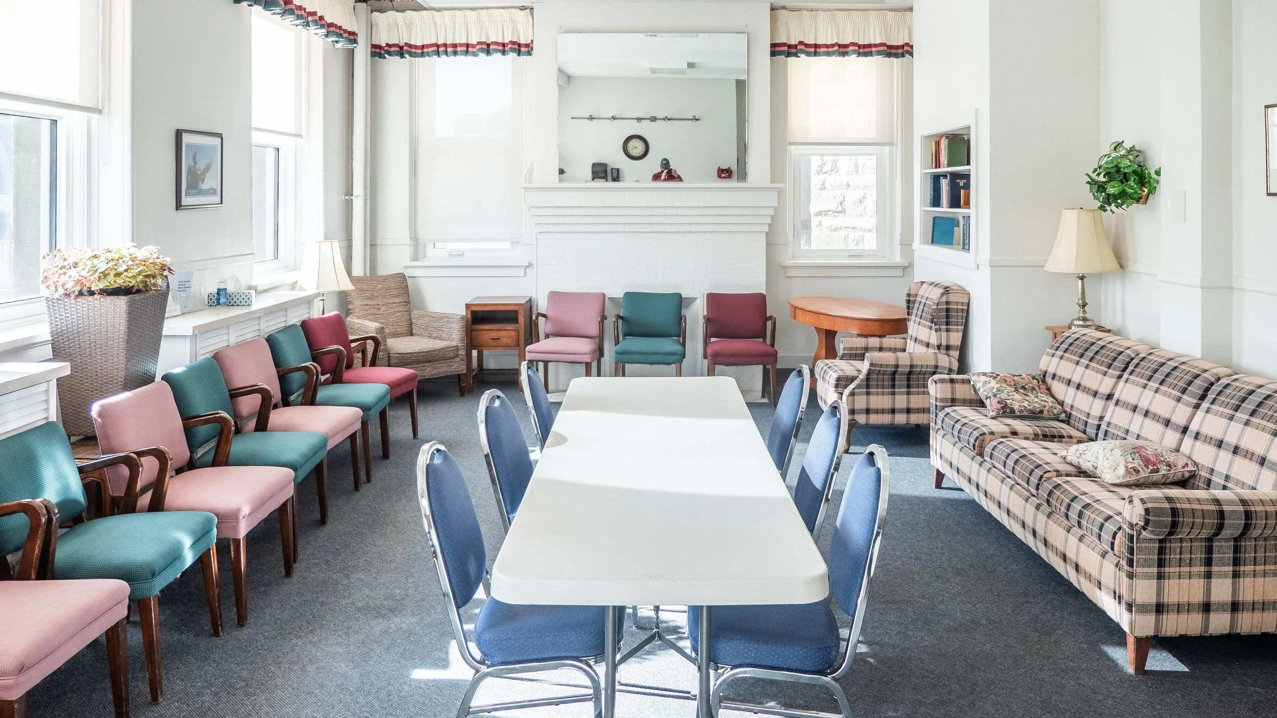 A sunlit room with a white rectangular table surrounded by blue chairs, a checkered sofa with pillows, and various other chairs. There's a fireplace with a mirror above it, a bookshelf, and lamps, creating a cozy, vintage atmosphere.