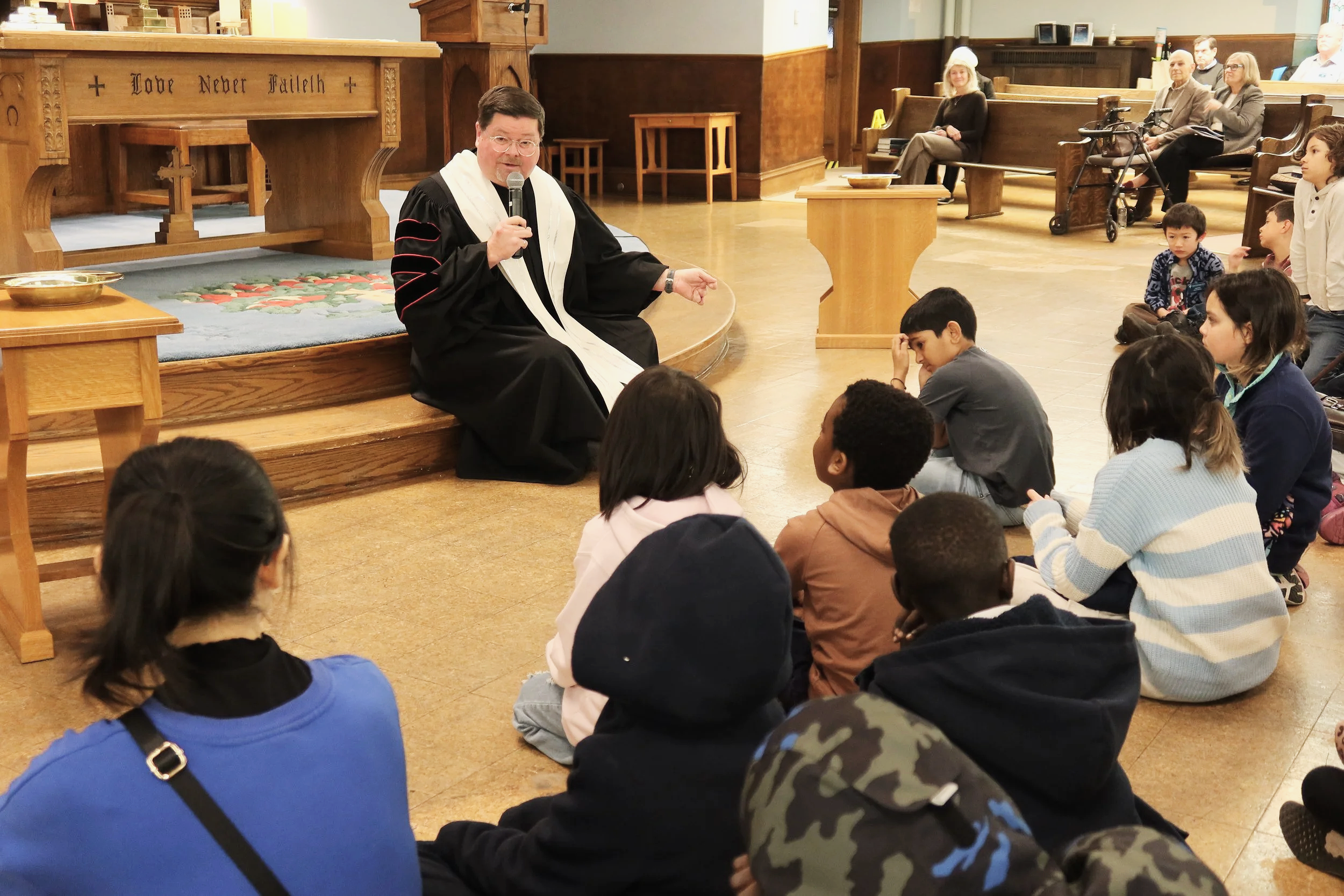 The Rev. Chris Joiner is sitting on a small stage in a church, speaking into a microphone to a group of children sitting on the floor in front of him, with some adults seated in the background. The Re
