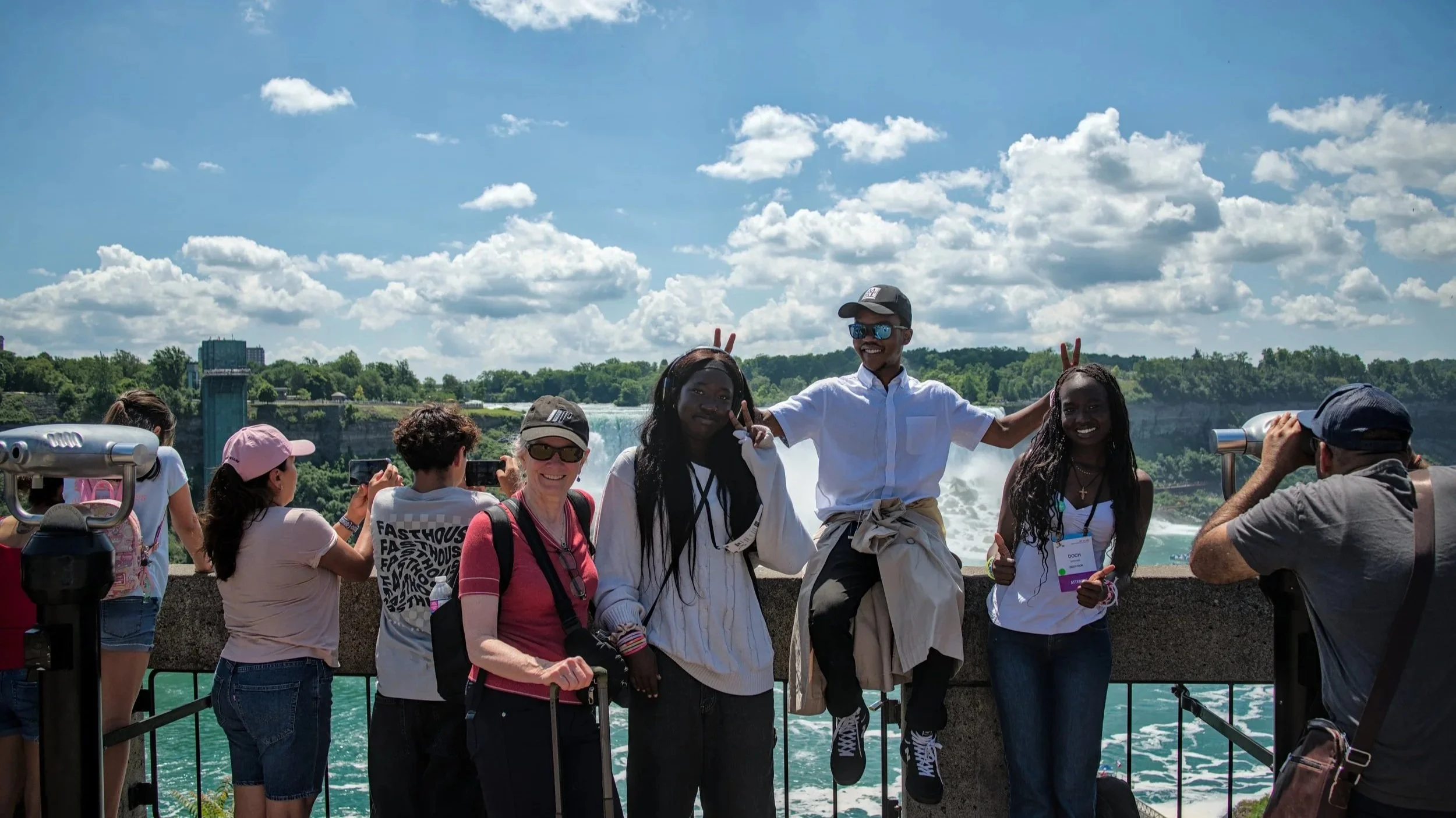 A group of youth taking photos at Niagara Falls, with the waterfall in the background and a clear blue sky with clouds.