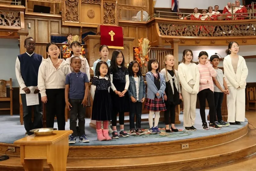 Children singing in a church choir, standing on a wooden stage with a wooden railing and choir members seated above.