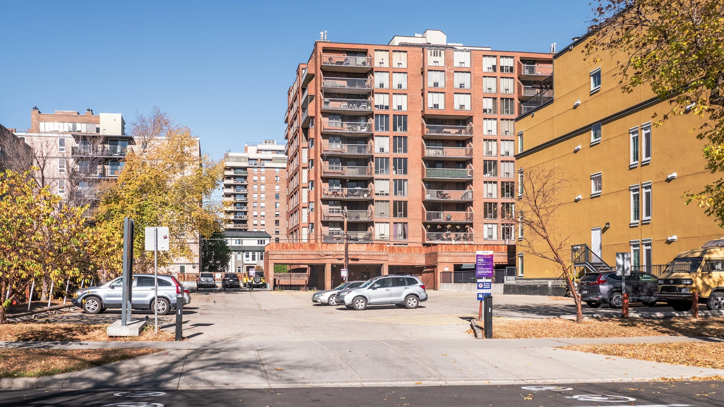 Urban parking lot with parked cars in front of multi-story residential buildings, some trees with autumn foliage, and a clear blue sky.