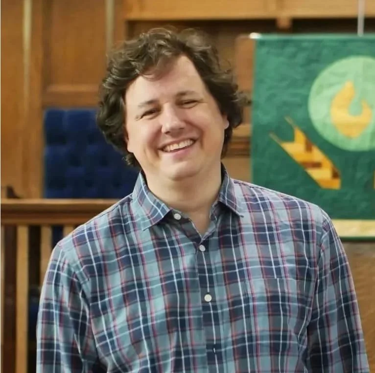 A smiling man with curly brown hair wearing a plaid shirt, standing indoors with wooden paneling and a green flag in the background.