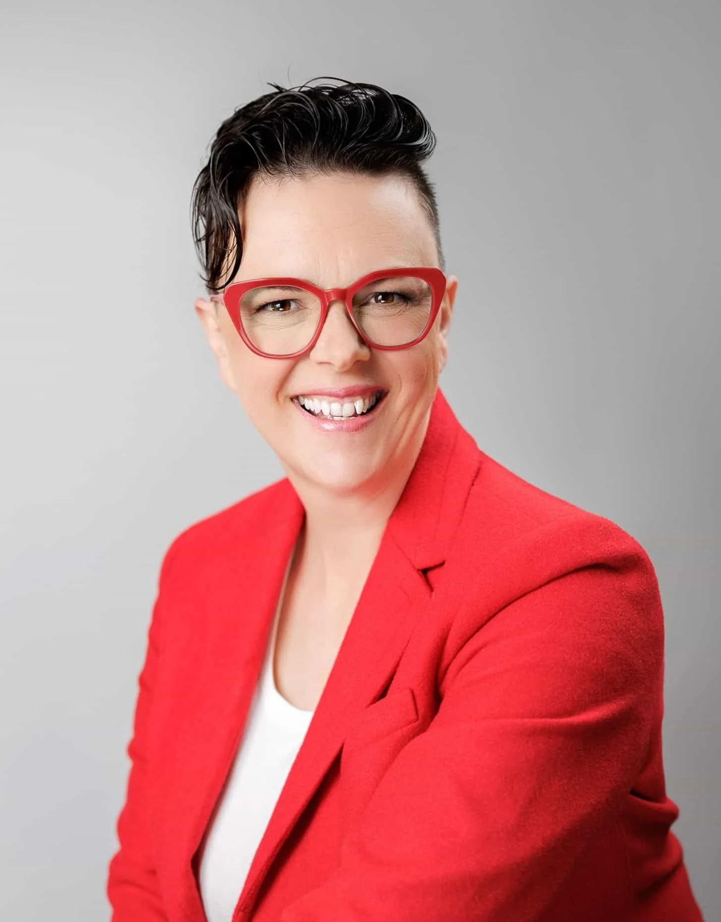 A woman smiling, wearing red glasses and a red blazer, against a neutral gray background.