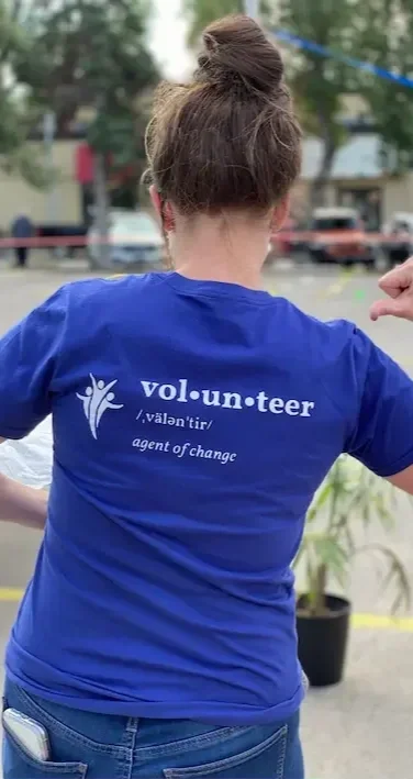 Back of a woman wearing a blue volunteer t-shirt with the words "vol•un•teer" and a logo, standing outdoors in a park.
