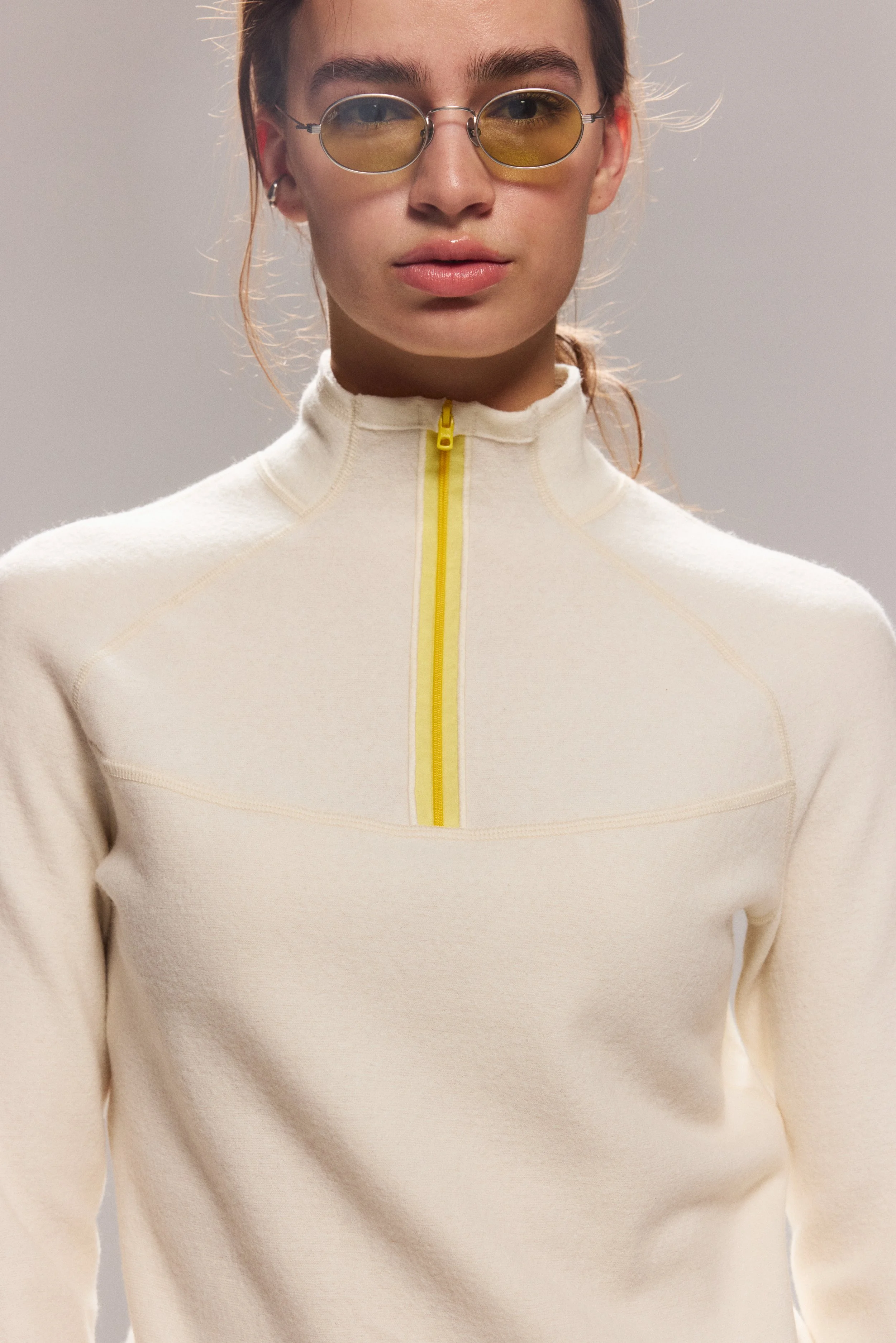 A person wearing a cream-colored high-neck athletic top with a yellow zipper in the front. They have on round yellow-tinted sunglasses and silver earrings, and their hair is tied back.