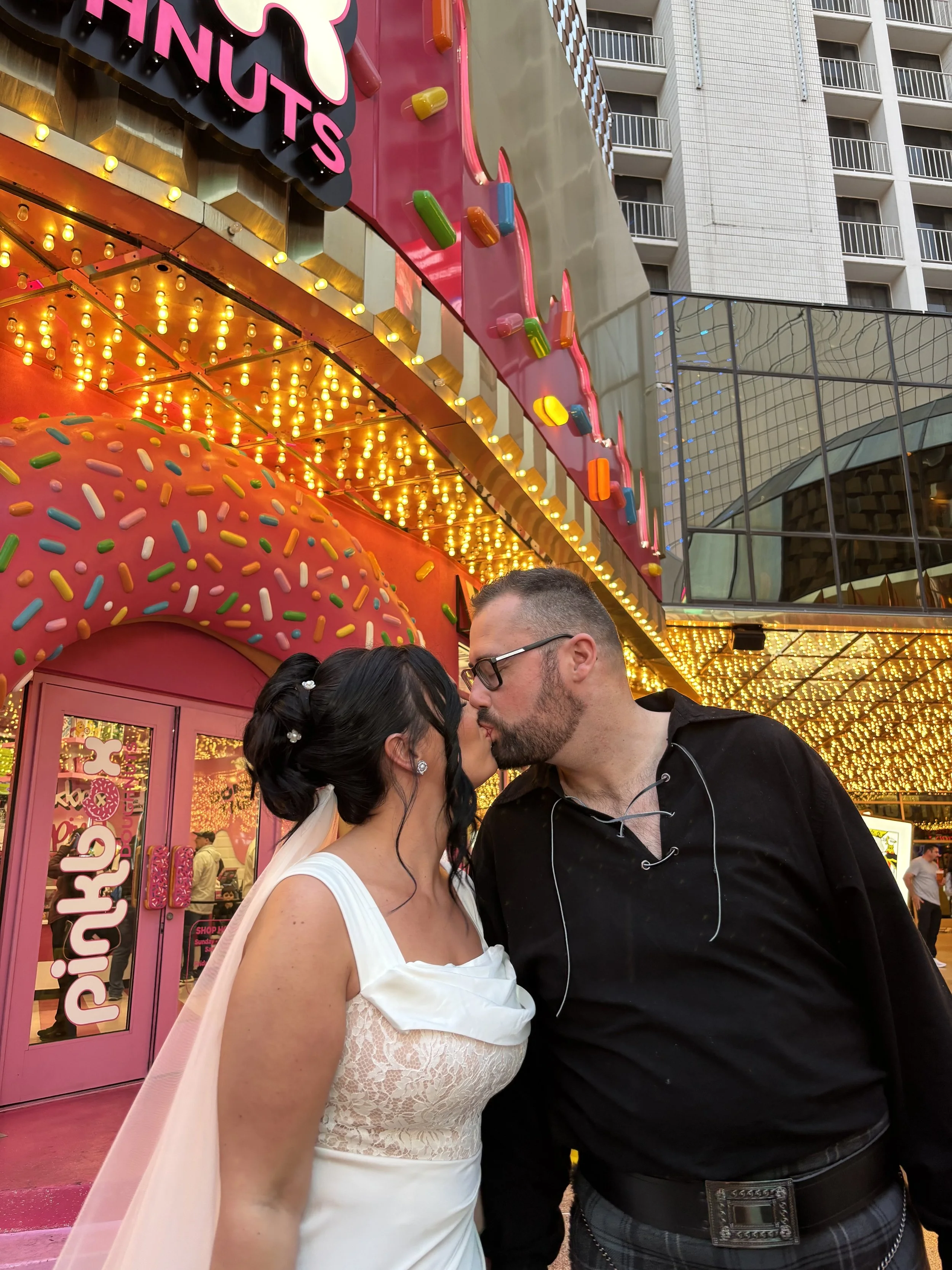 A couple kissing outside of a bright pink shop decorated with colorful sprinkles and bright lights, with a pink cupcake-shaped archway. The woman wears a white wedding dress, and the man wears a black shirt and glasses.