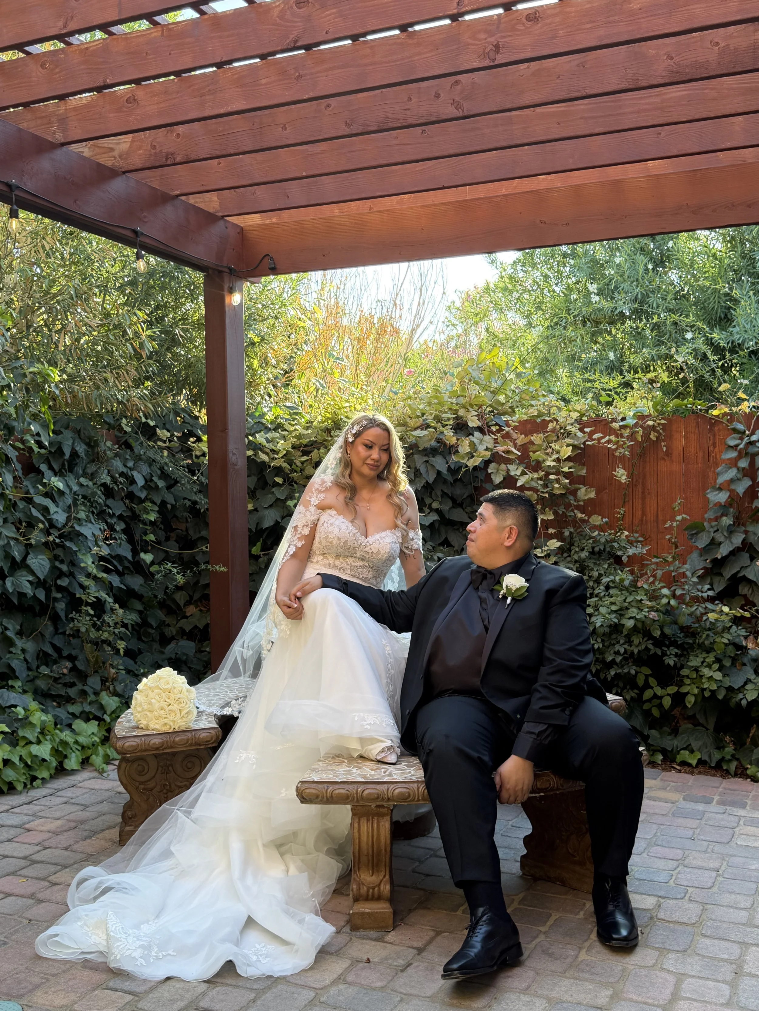 Bride in a wedding dress sitting on a bench, holding hands with groom in a black tuxedo, outdoors under a wooden pergola with greenery in the background.