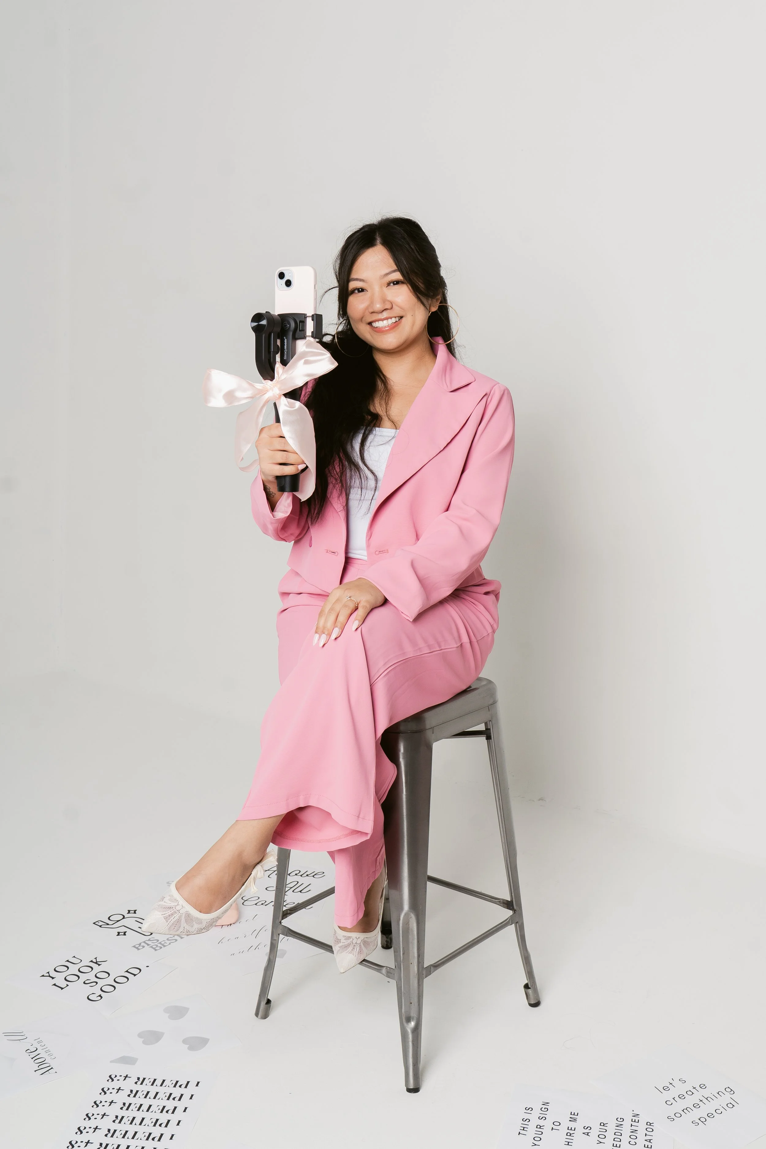 A woman with long dark hair, wearing a pink suit and white shoes, sitting on a tall metal chair against a plain white background. She is smiling, holding a selfie stick with a bow on it, and taking a photo.