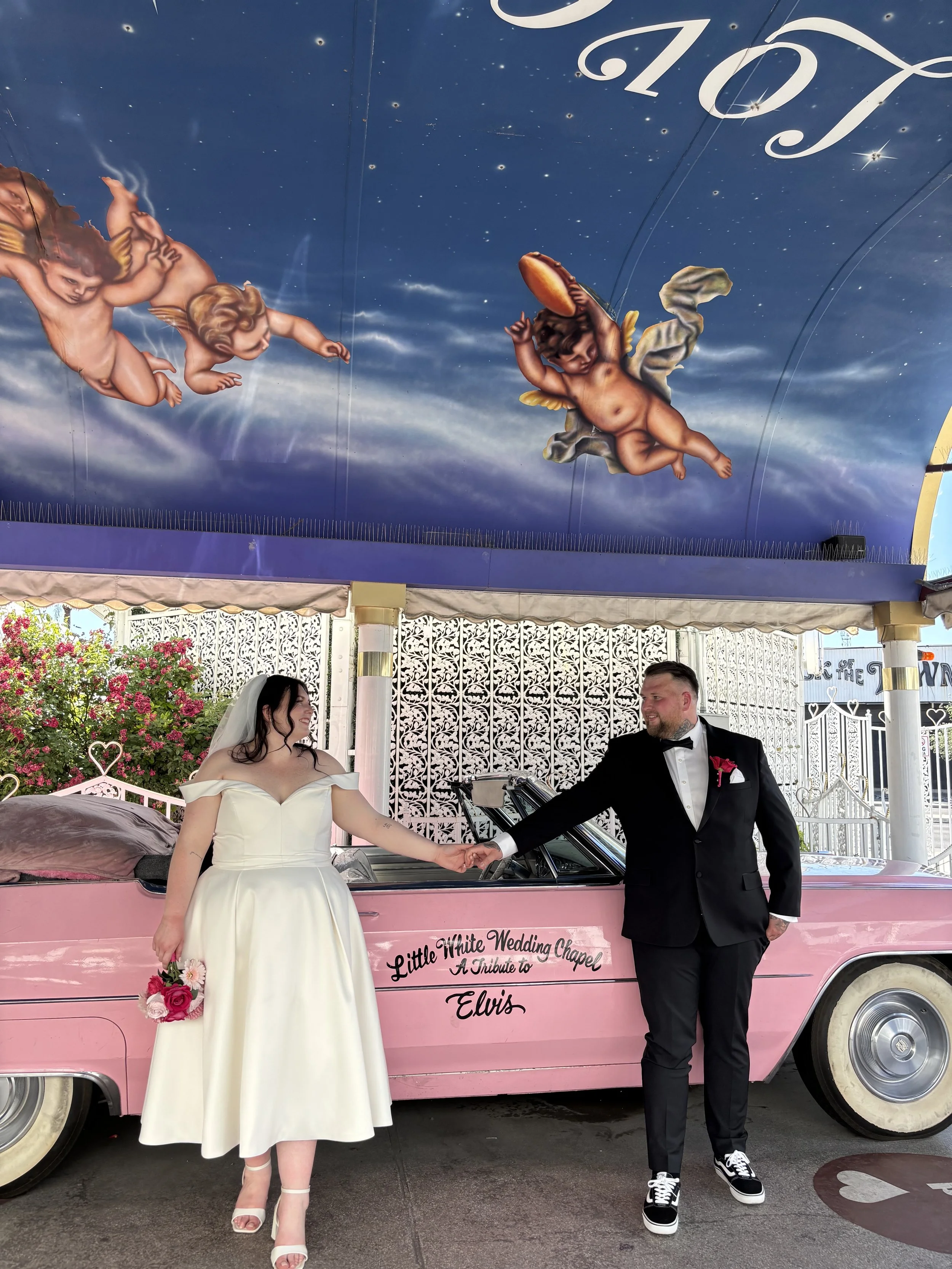 A bride and groom holding hands in front of a pink vintage car with a sign that reads "Little White Wedding Chapel A Tribute to Elvis". The ceiling above features classic Christian artwork with cherubs and clouds.