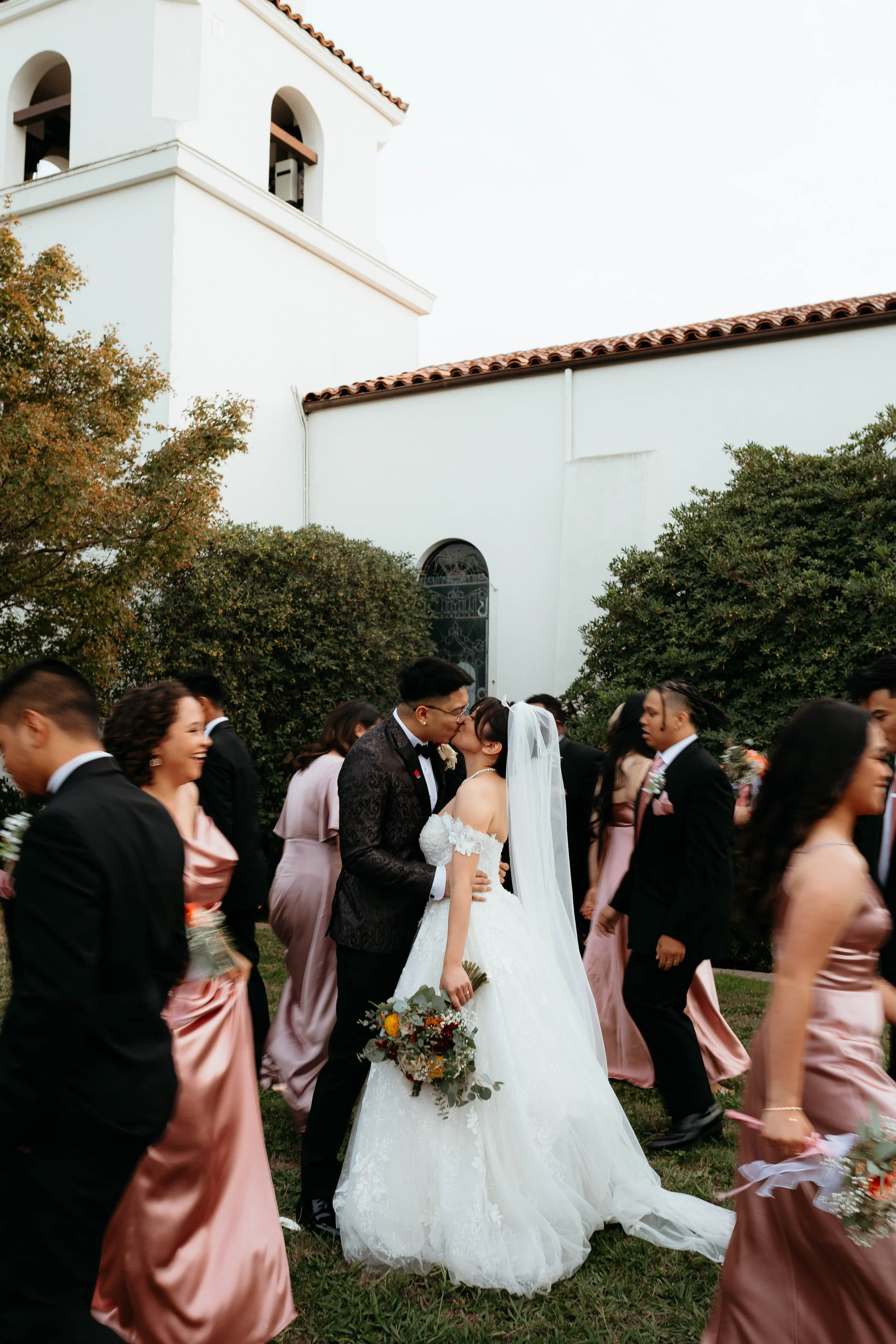 Bride and groom kiss at their wedding surrounded by wedding guests outdoors with a white building and greenery in the background.