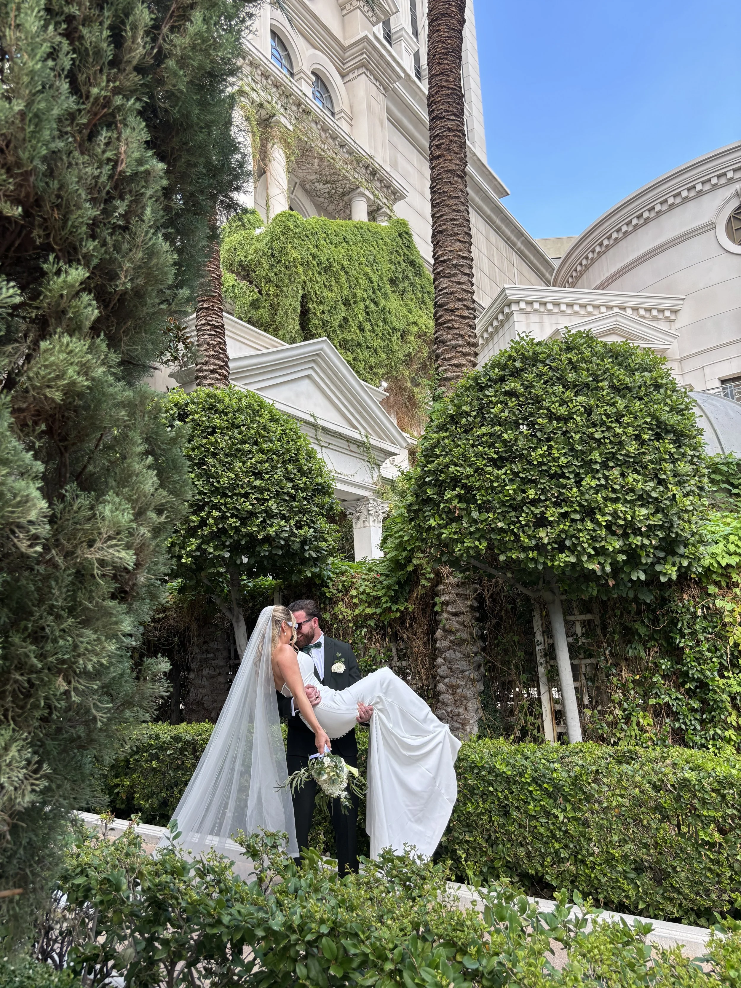 A newlywed couple, the groom in a black tuxedo and the bride in a white wedding dress with a veil, sharing a kiss outdoors surrounded by lush greenery and tall trees, with a white building in the background.
