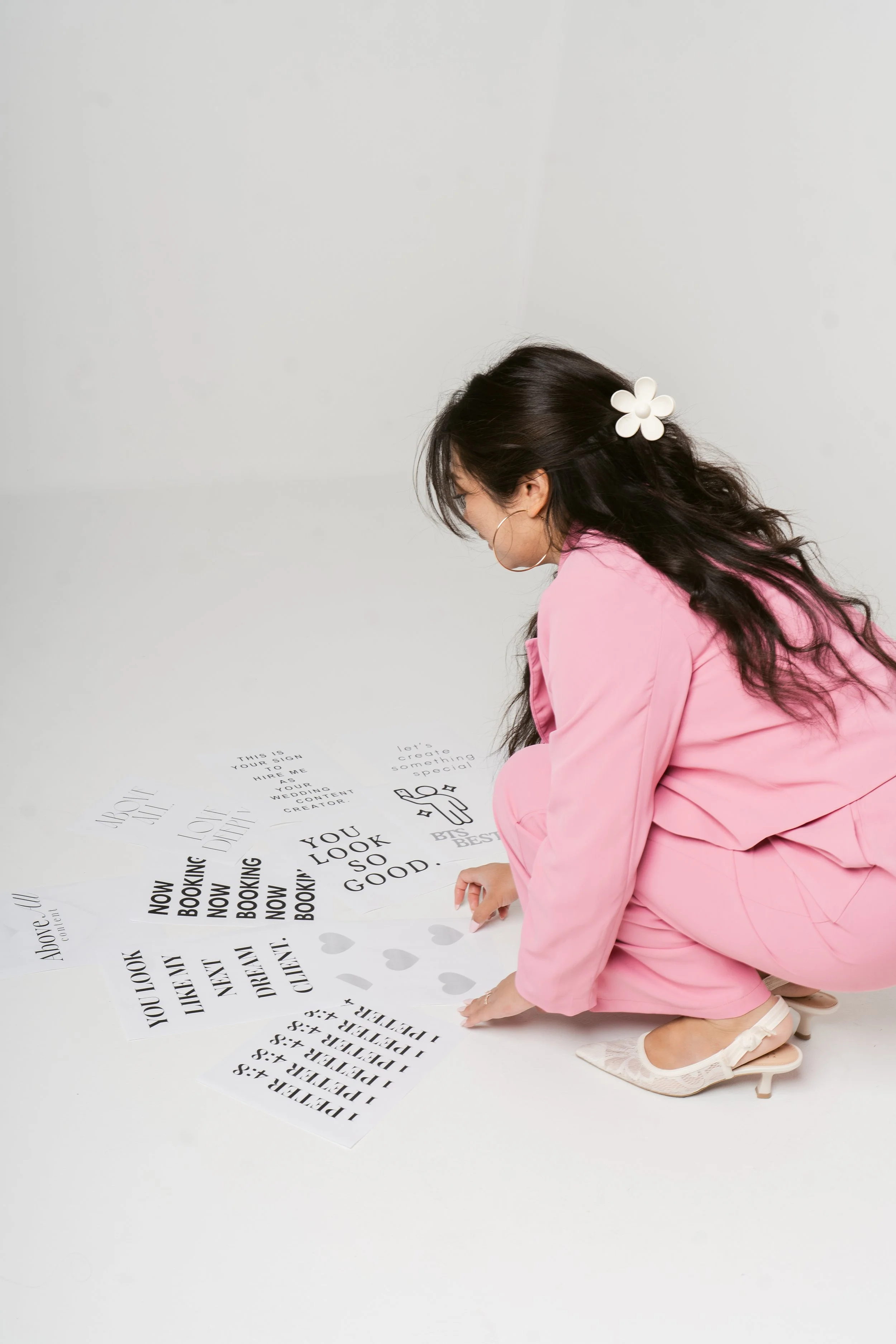 Woman in pink suit crouching on white floor, arranging printed posters with black text and designs.