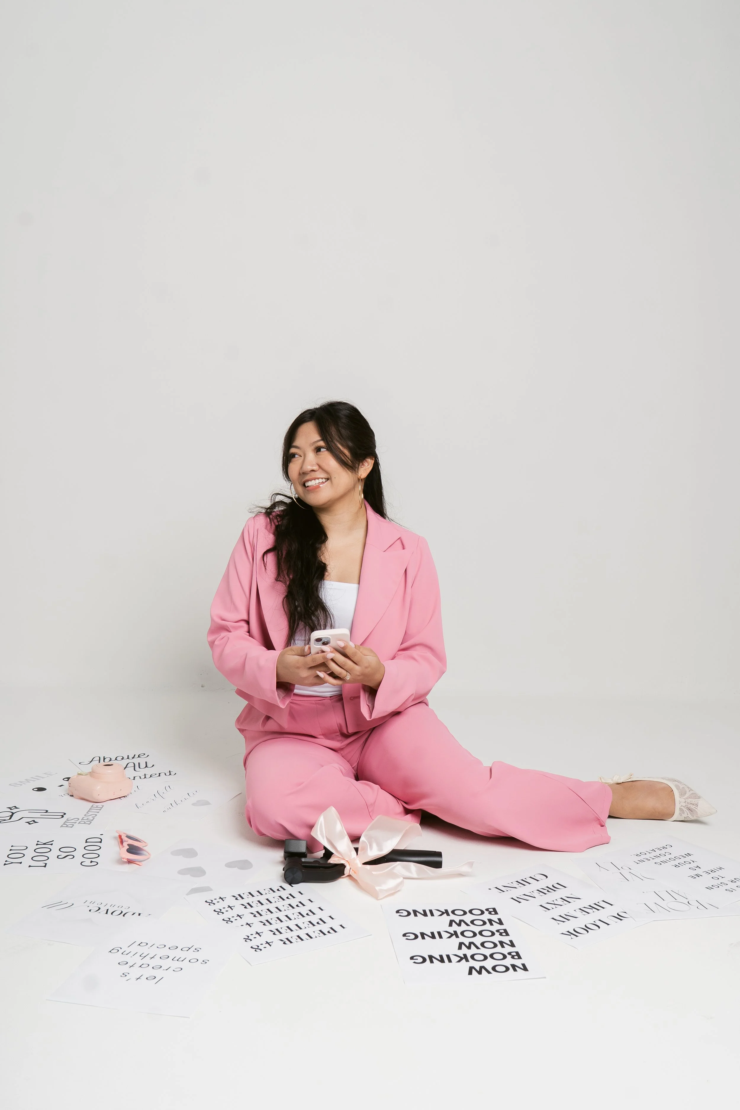 A woman in pink pants and blazer sitting on the floor, looking to her left and smiling, holding a phone, surrounded by papers, a pink piggy bank, a ribbon, and frame glasses, against a plain white background.