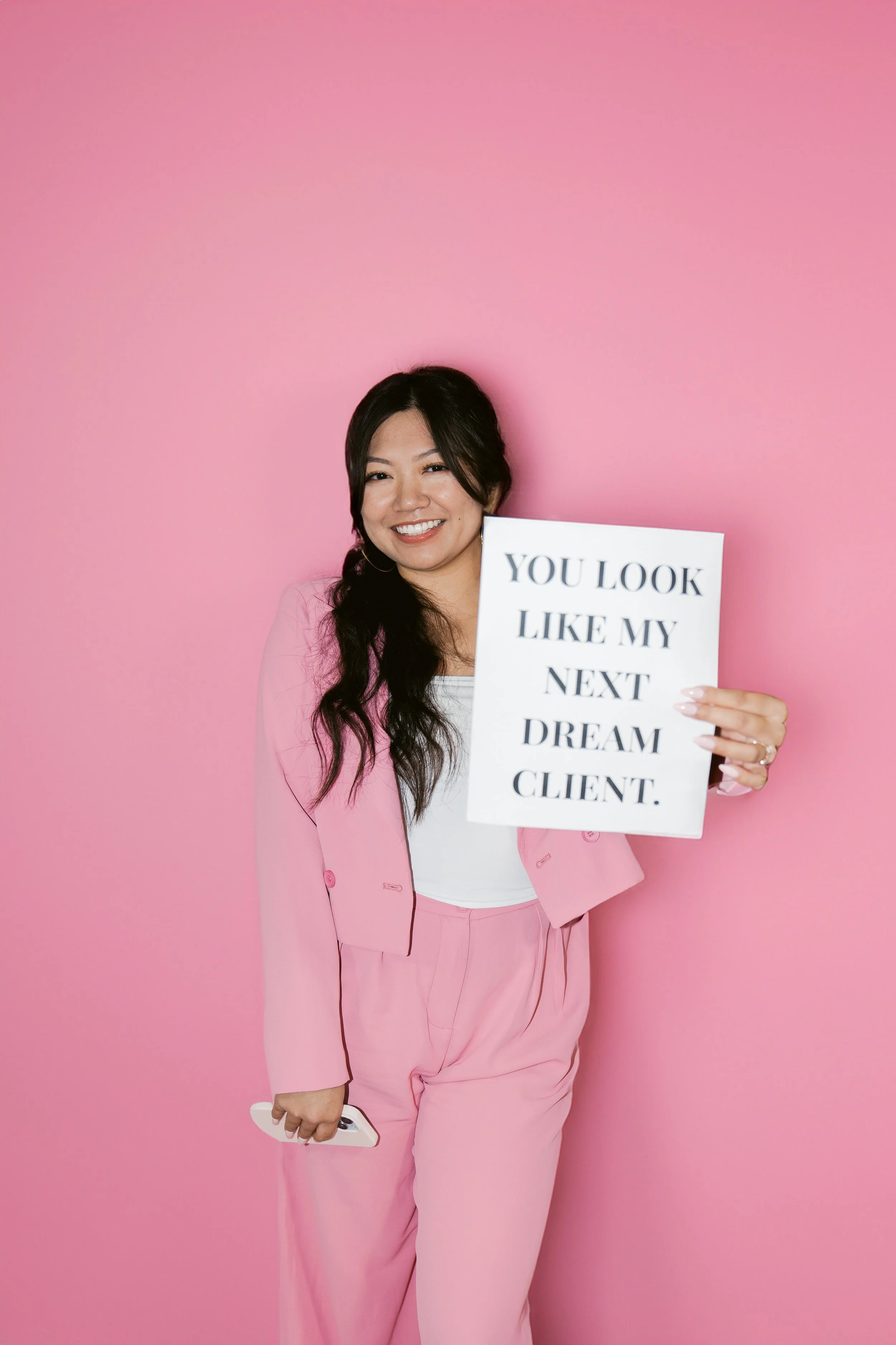 A woman with long dark hair, dressed in pink suit and white top, smiling and holding a sign that says 'You look like my next dream client,' against a pink background.