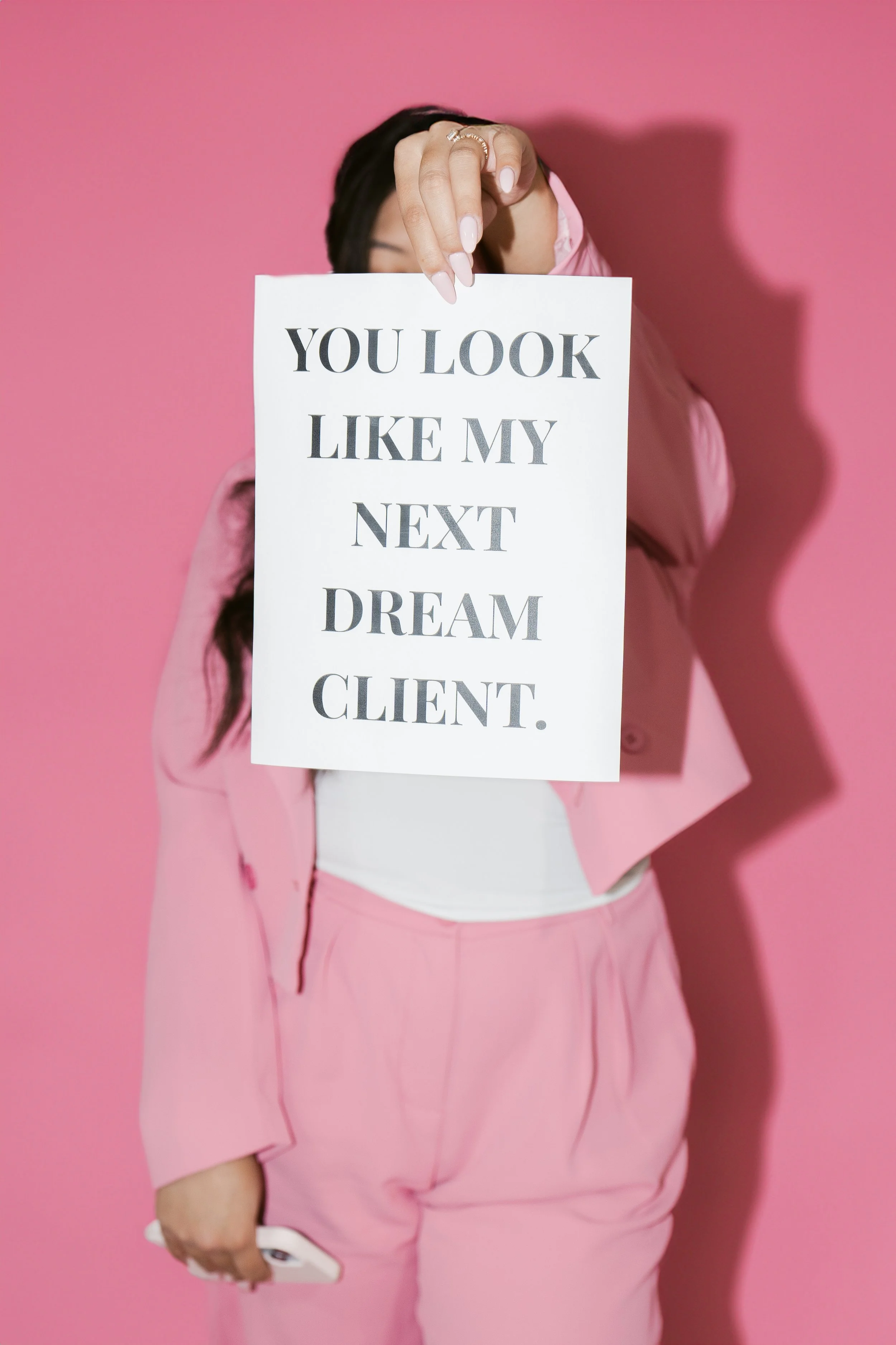 A woman in pink outfit holding a sign that says "You look like my next dream client" in front of a pink background.