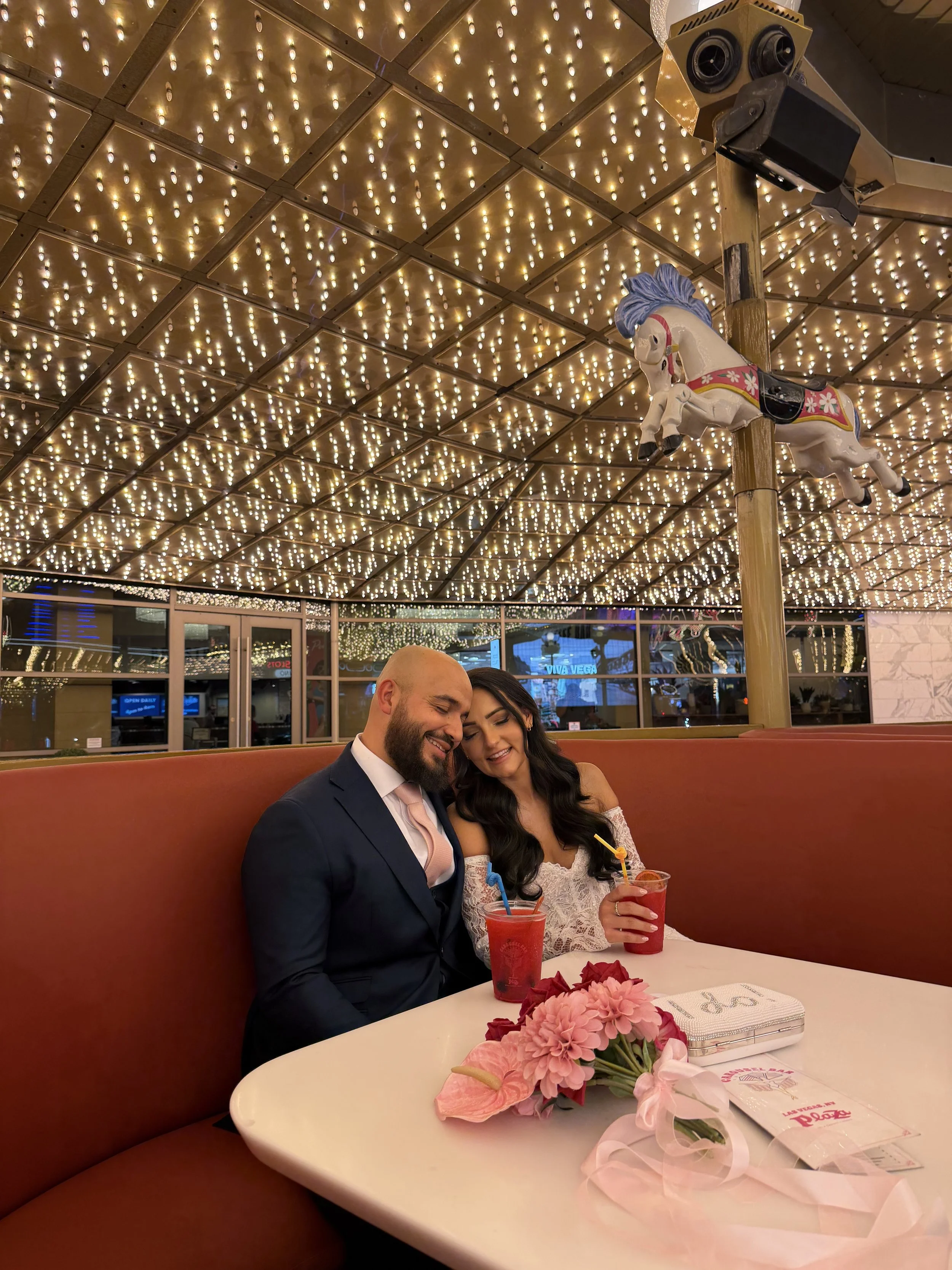A couple sitting together at a table in a decorated restaurant with a ceiling covered in string lights. The man has a beard and is dressed in a dark suit, and the woman has long dark hair and is wearing a white lace dress. They are smiling and holding drinks with colorful straws. A bouquet of pink flowers and some papers are on the table. There is a carousel horse hanging from a pole above them.