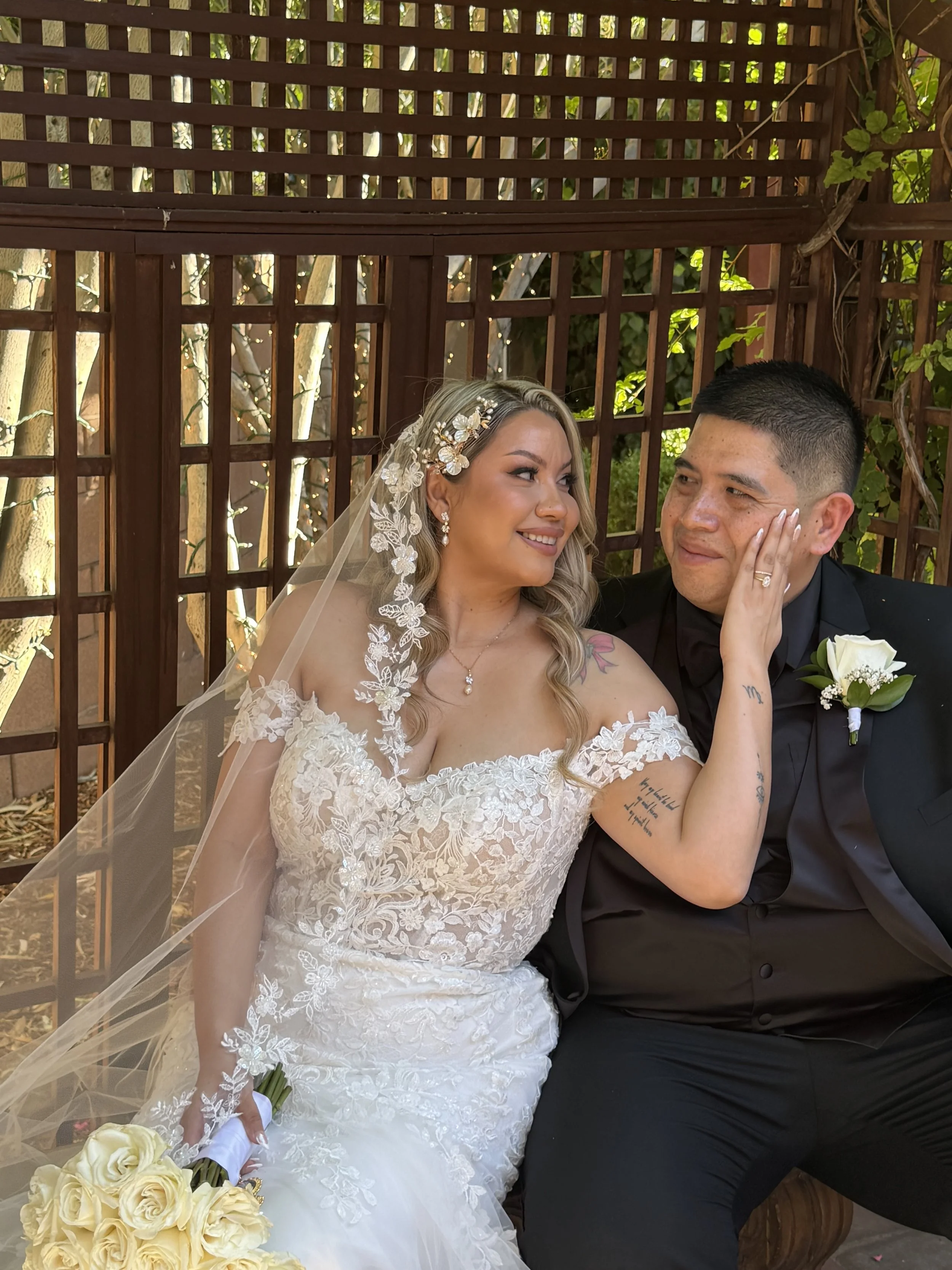 Bride in a lace wedding gown with a veil and floral headpiece, holding a bouquet of white roses, sitting next to groom in a black tuxedo with a white boutonniere, both smiling and gazing at each other, indoors with wooden lattice backdrop.