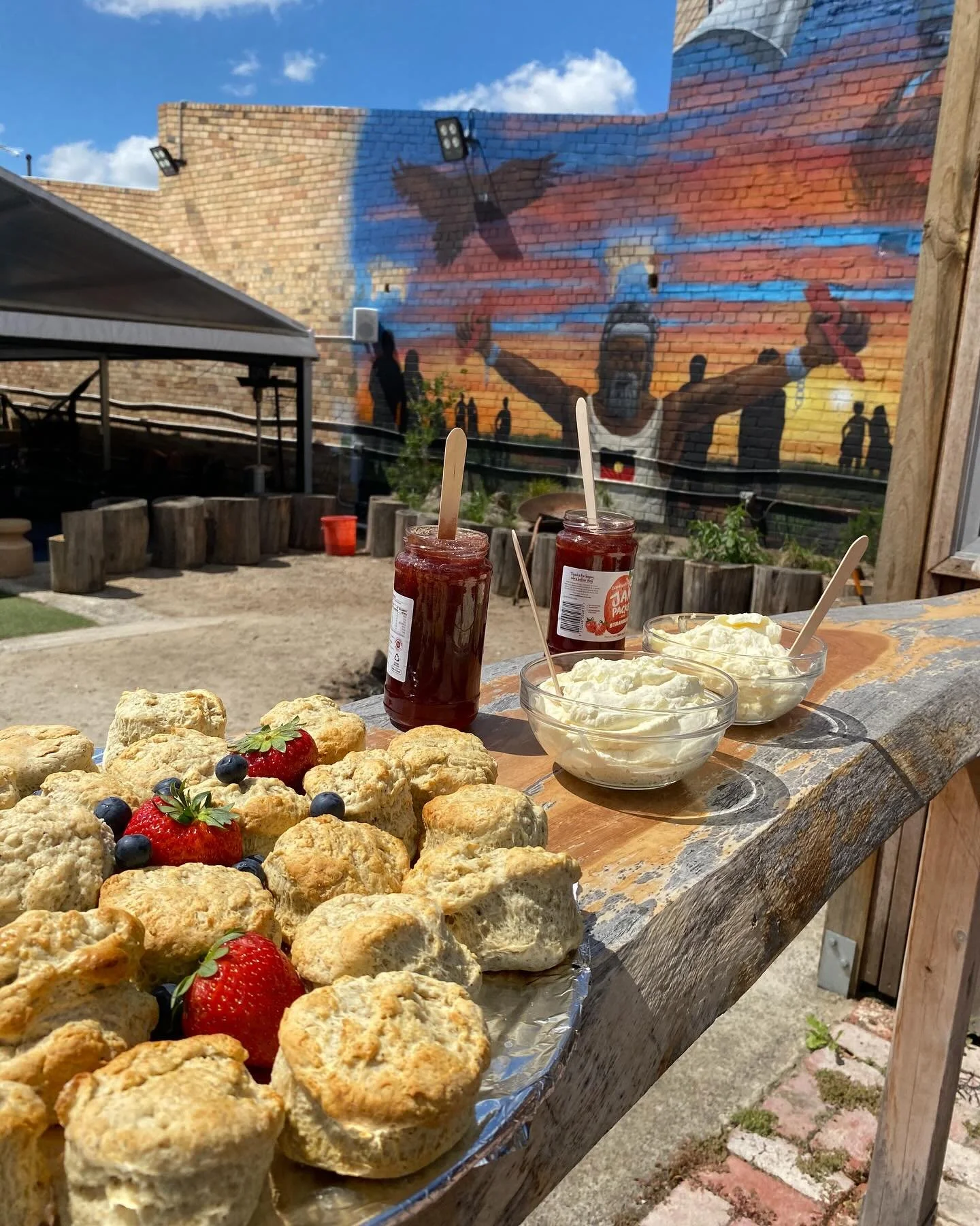 Wooden table with scones topped with strawberries and blueberries, jars of strawberry jam, and bowls of whipped cream set outdoors under blue sky with a brick wall mural of people and a large bird in the background.