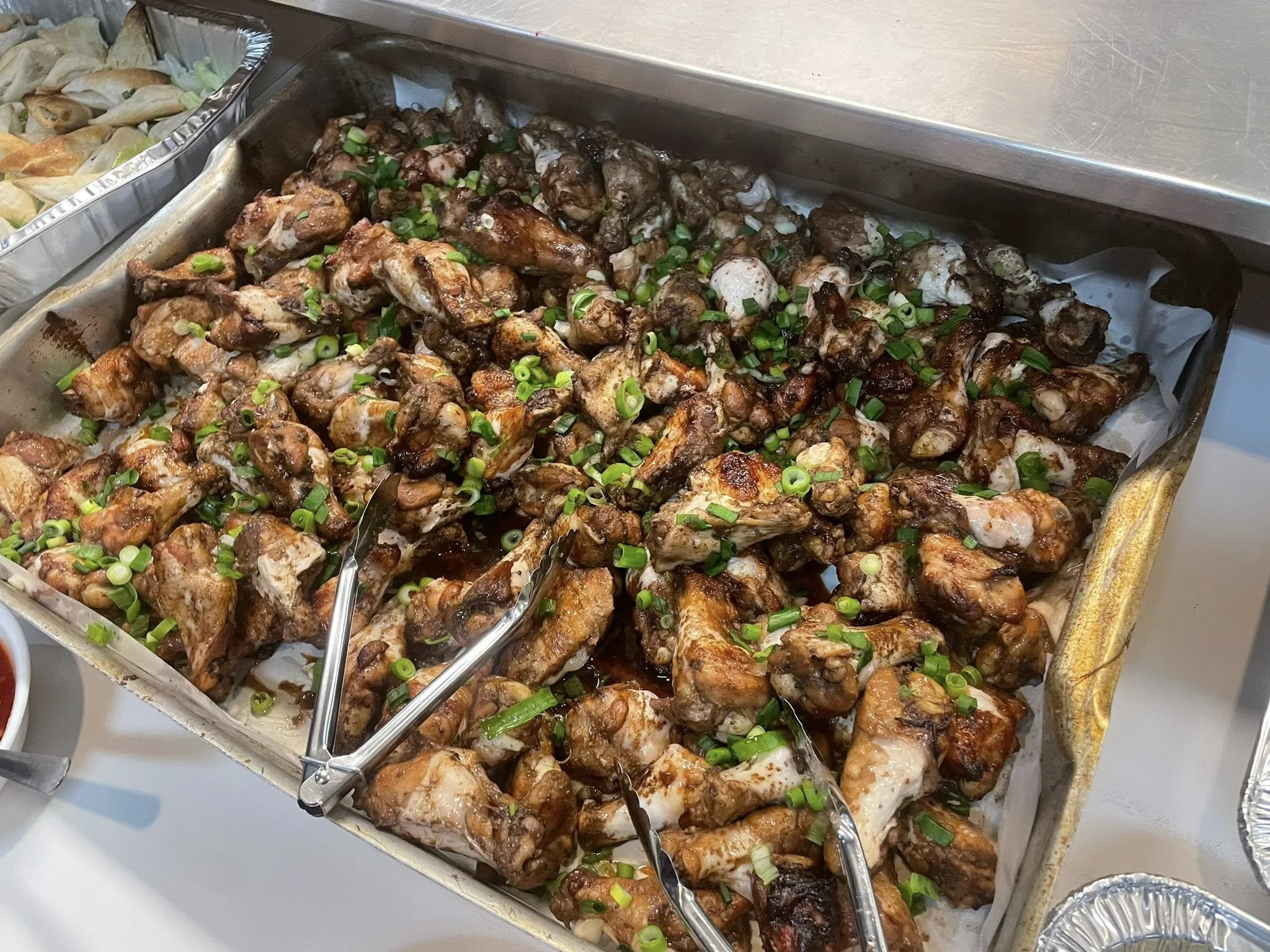 Tray of cooked chicken wings topped with chopped green onions, served with tongs for serving.