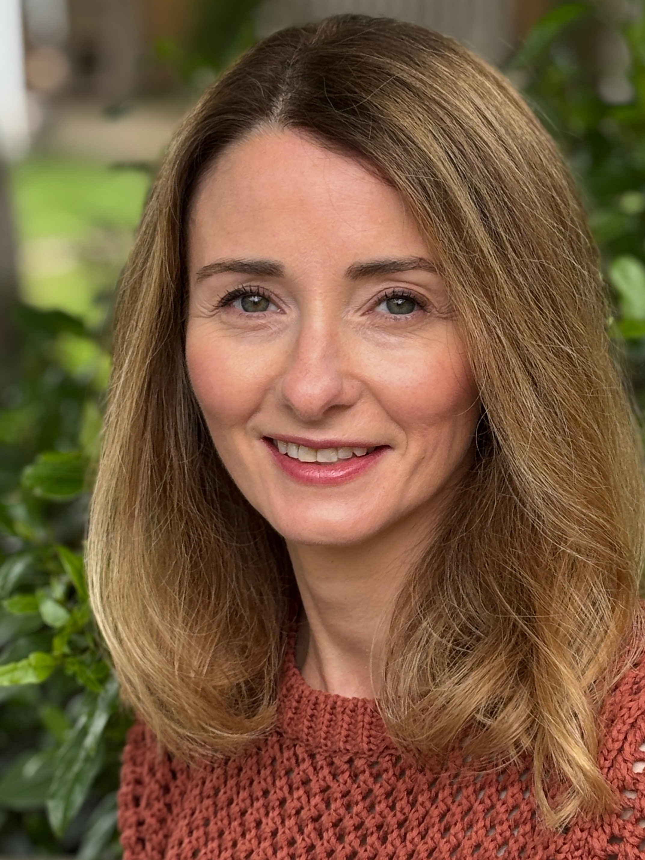 Close-up of a woman with light brown hair, green eyes, smiling, wearing a red knitted sweater, with green foliage in the background.