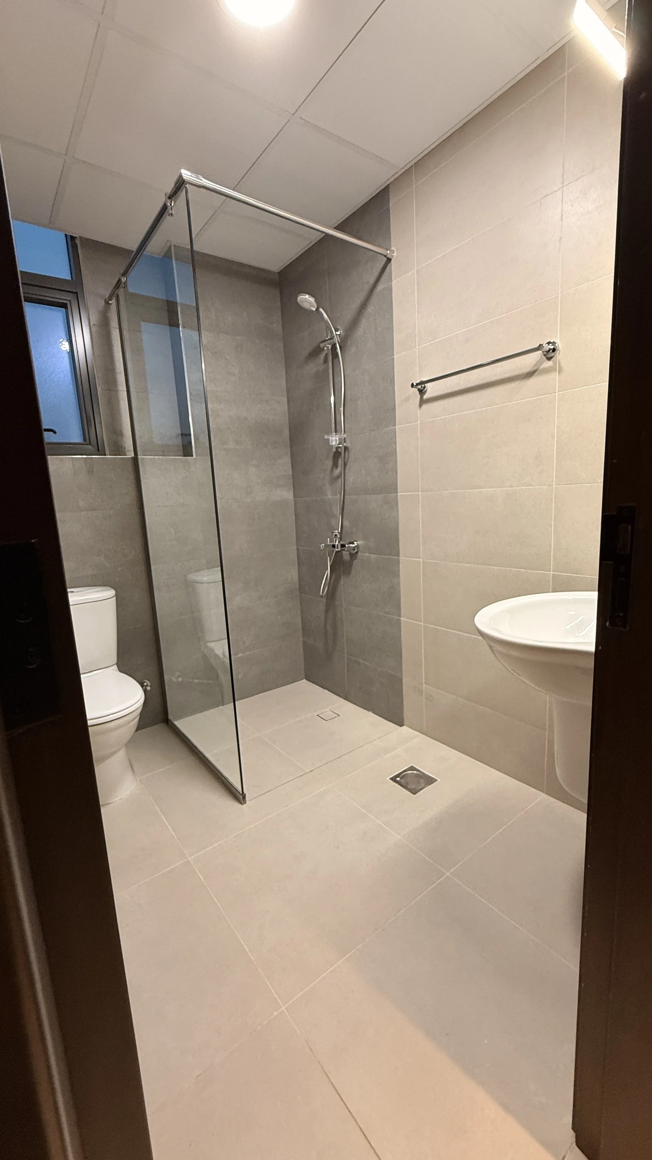 A modern bathroom with beige tiles, a walk-in shower with gray accent wall, a glass enclosure, a ceiling light, a small window, a white toilet, a wall-mounted white sink, .residences oman, freehold, apartments visa residency muscat residences in duqm
