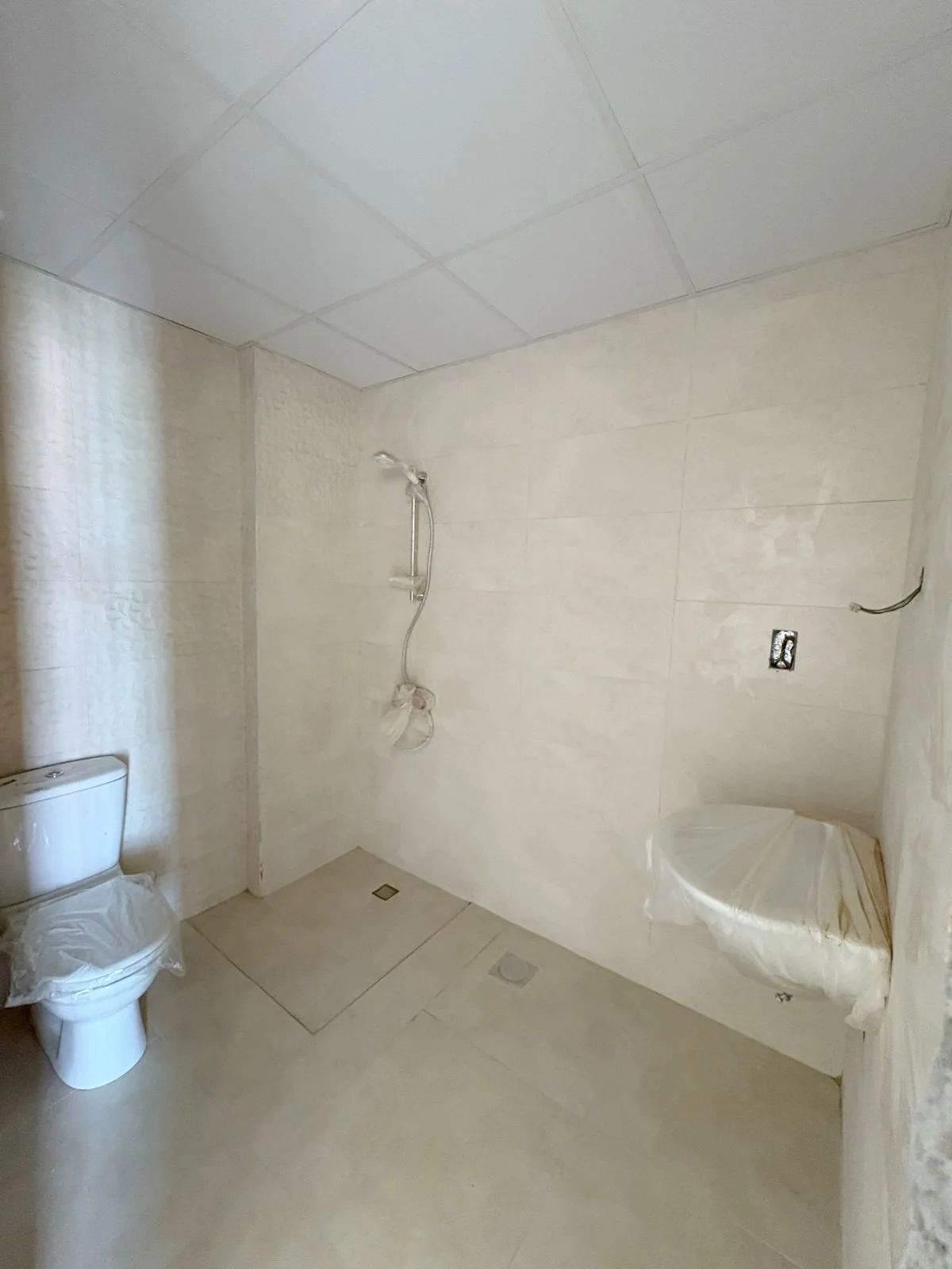 Unfinished bathroom with a toilet in the lower left corner, a showerhead on the wall, missing fixtures, and plastic covering some parts of the walls. Residences Oman Duqm