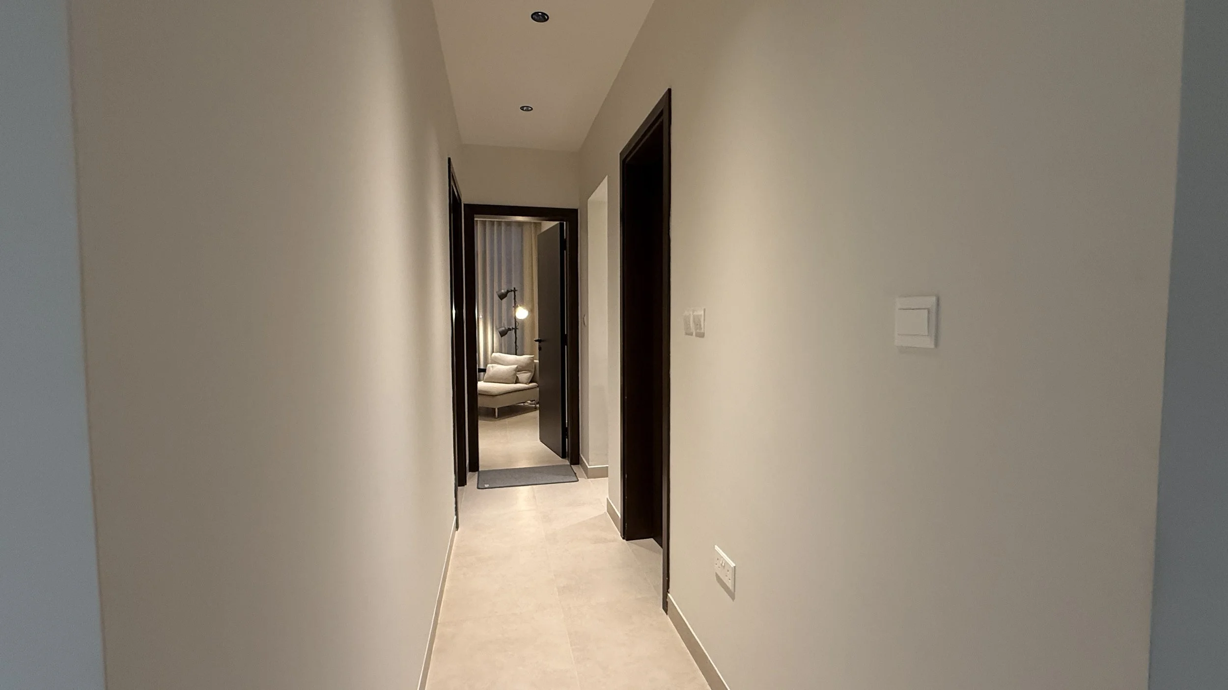 A hallway in a modern apartment or hotel with beige tile flooring, cream-colored walls, and dark wood door frames. At the end, a room with a beige sofa, .residences oman, freehold, apartments visa residency muscat residences in duqm
