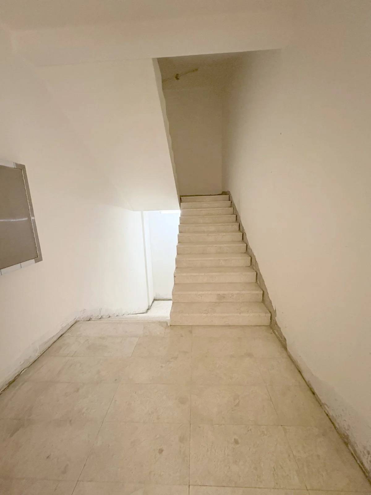Empty indoor staircase with beige tiled floor and off-white walls, leading up to a small landing and a door at the top. Residences Oman Duqm