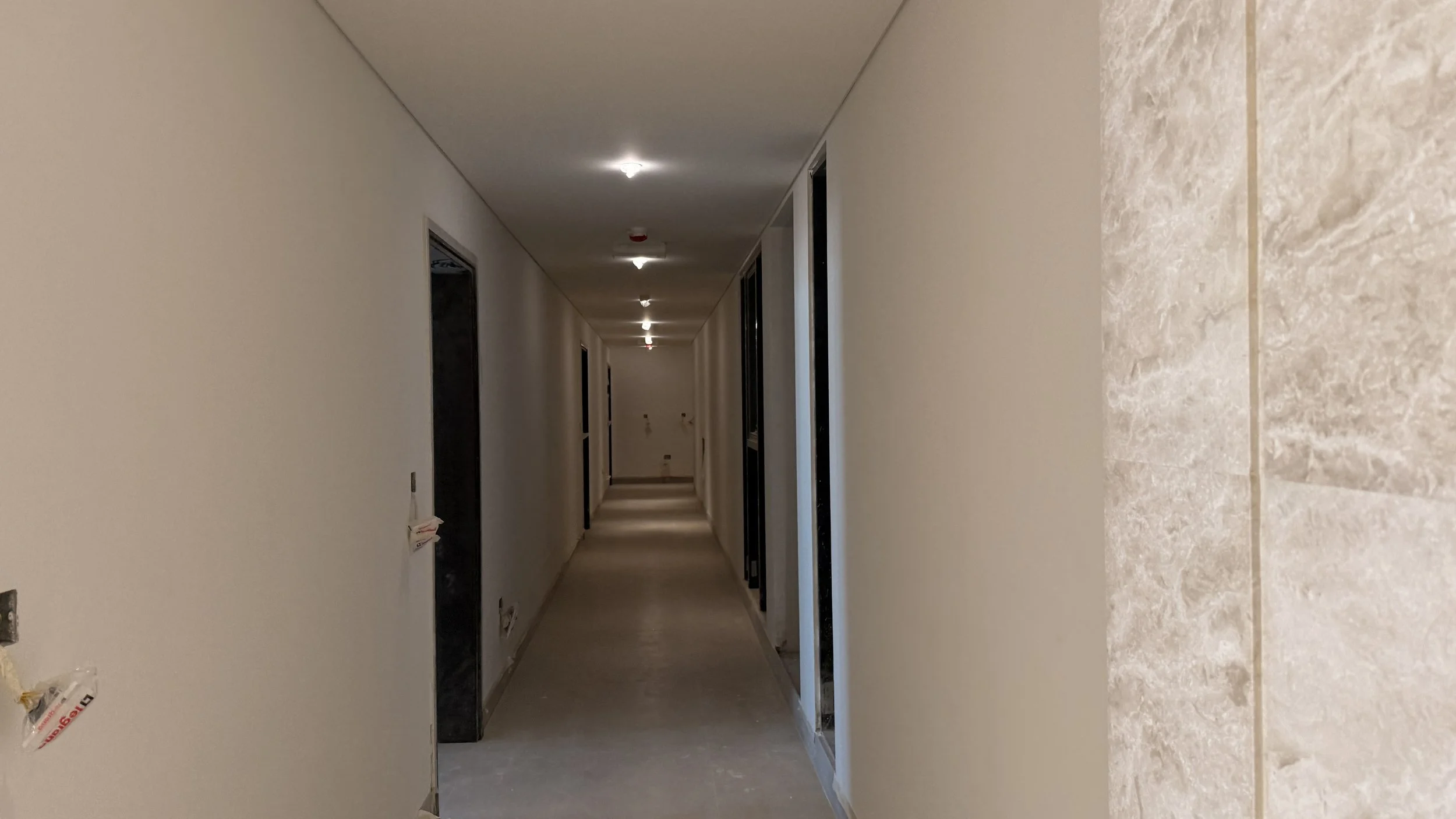 Long indoor hallway with beige walls and ceiling, scattered ceiling lights, and several doorways along the sides, under construction or renovation..residences oman, freehold, apartments visa residency muscat residences in duqm
