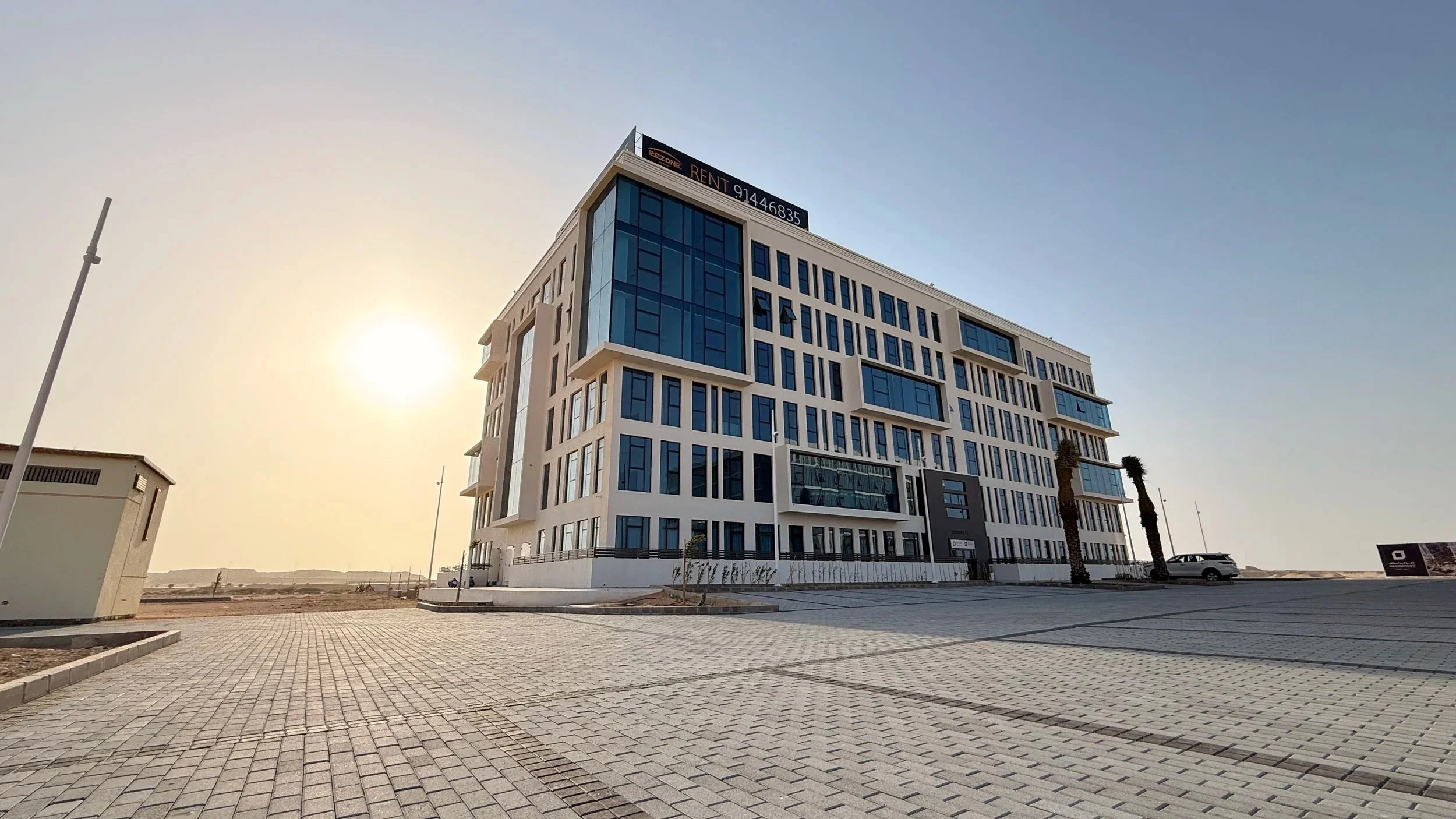 Modern multi-story building with blue glass windows and white exterior, under a clear sky with the sun low on the horizon.