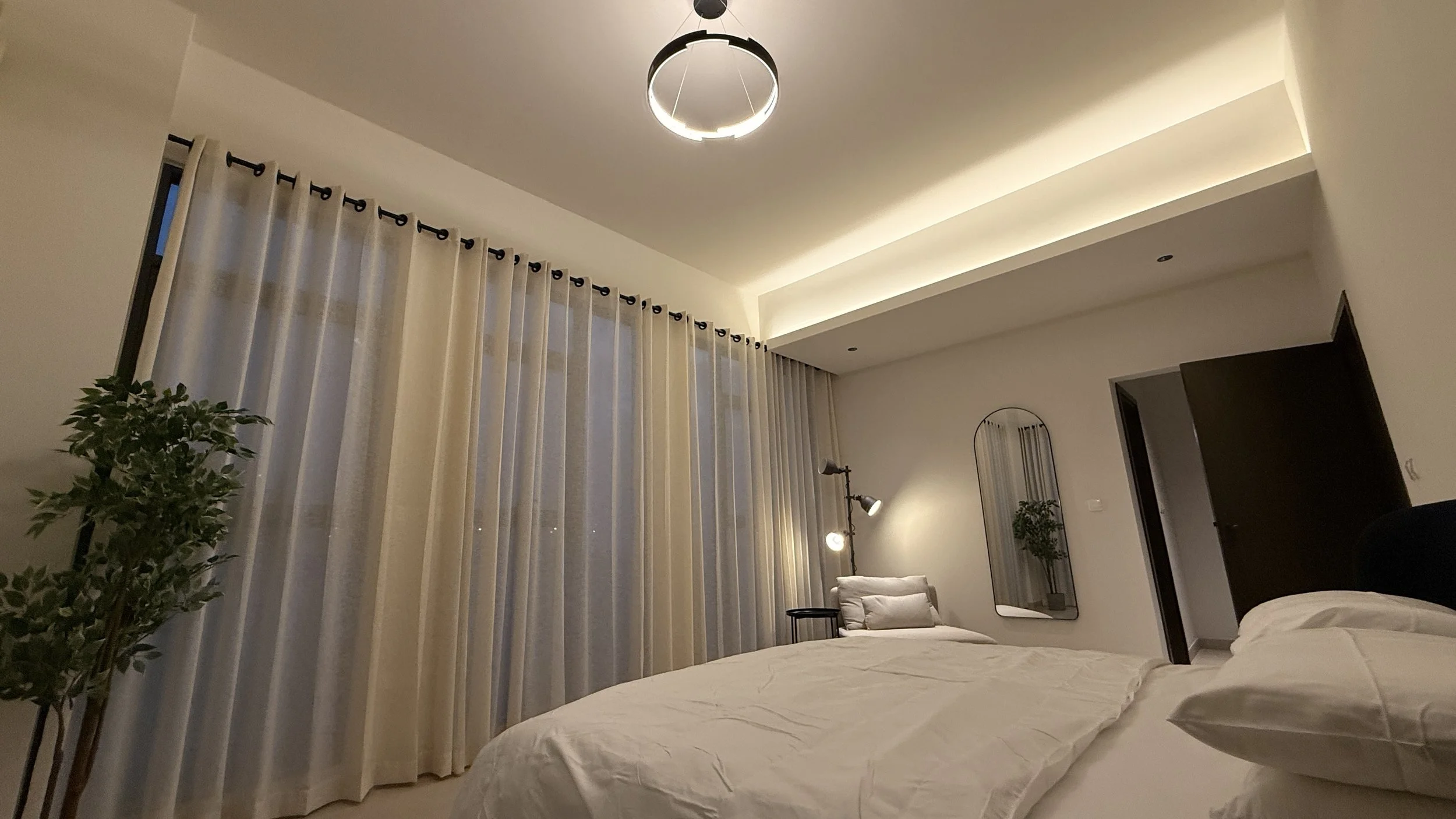 A modern bedroom with white walls and ceiling, beige curtains covering windows, a bed with white bedding, a black side table, and a standing lamp. .residences oman, freehold, apartments visa residency muscat residences in duqm