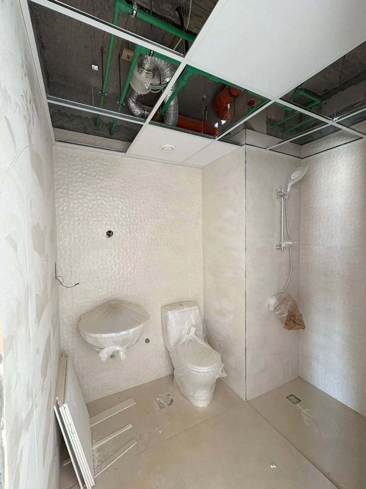 A bathroom under construction with a toilet and shower, ceiling panels removed revealing pipes and ductwork, walls covered with textured tile, and construction materials on the floor. Residences Oman Duqm
