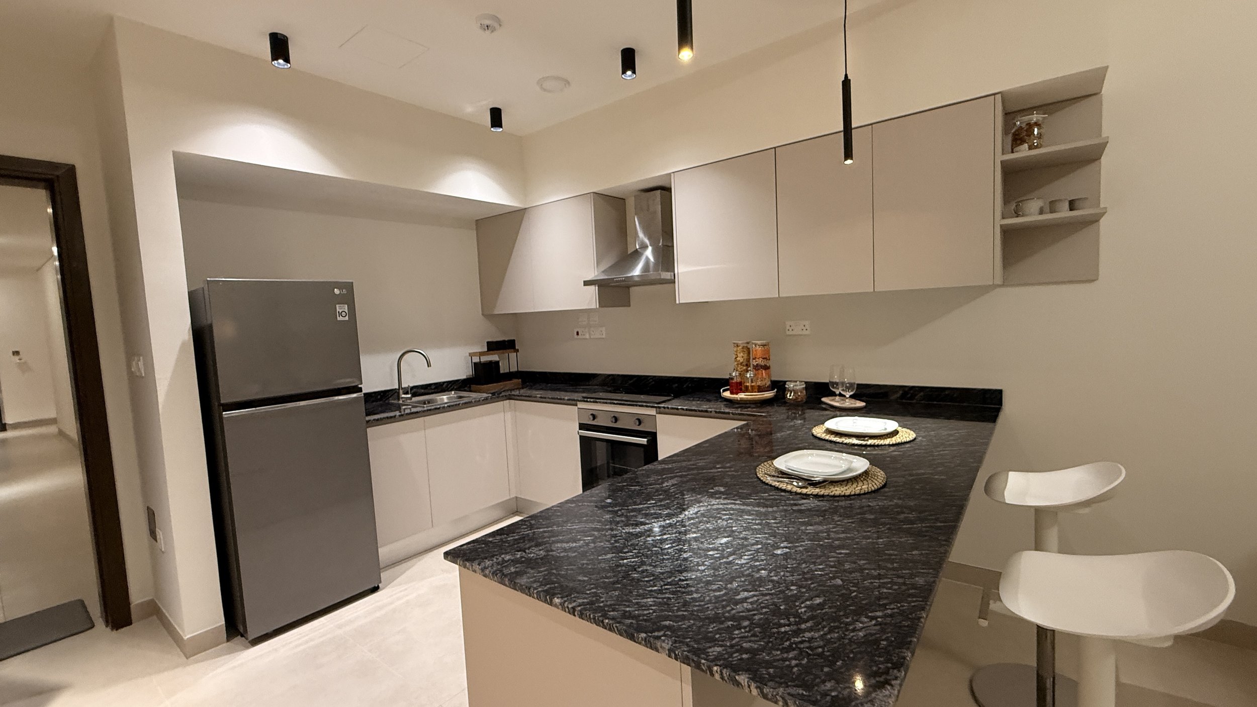 Modern kitchen with black granite countertops, white cabinetry, stainless steel refrigerator, and bar chairs. Decor includes plates and glasses on the counter..residences oman, freehold, apartments visa residency muscat residences in duqm