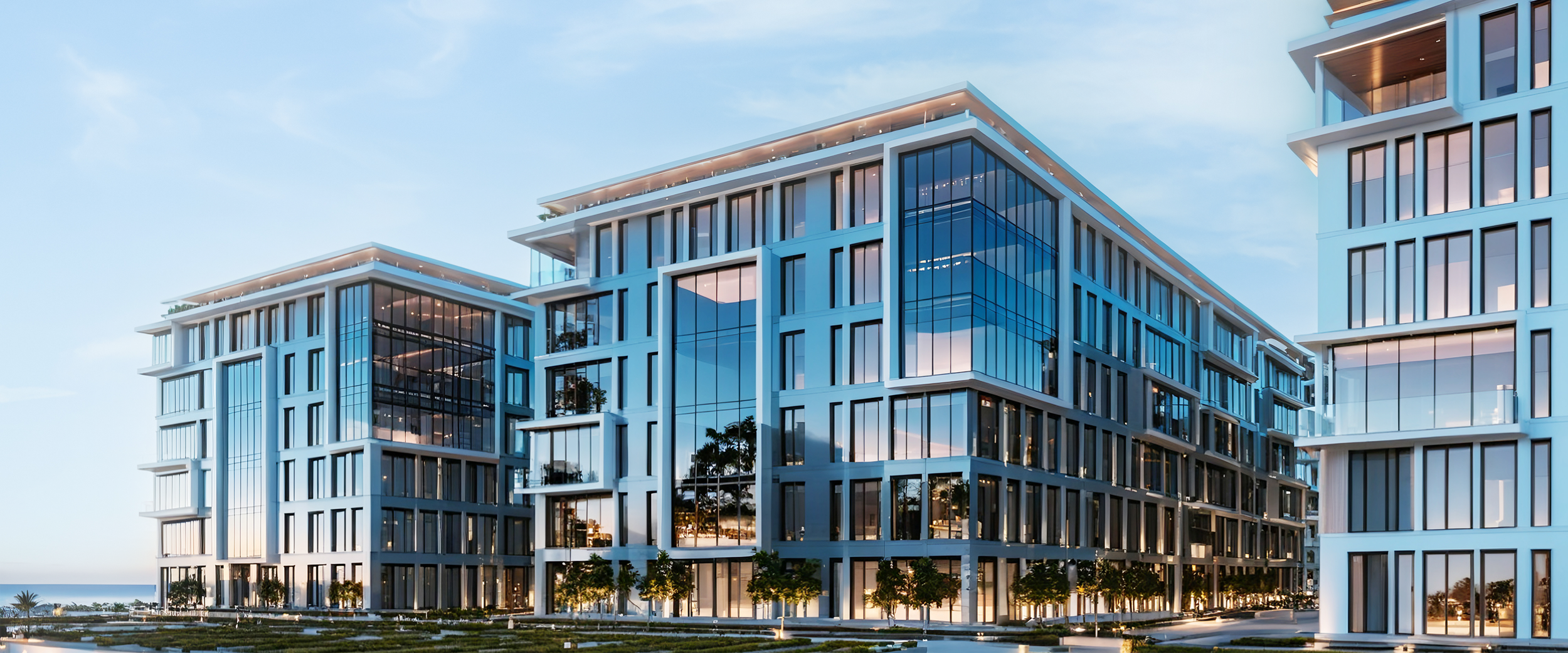 Modern multi-story glass building with white framing and rows of windows, trees at the base, and clear sky in the background.