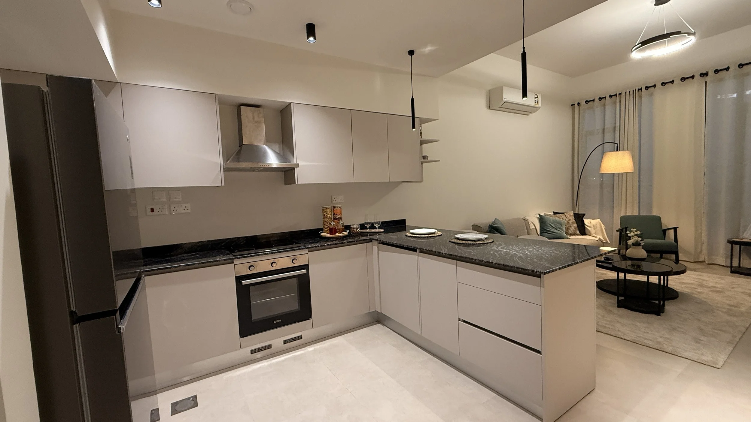 Modern kitchen with white cabinets, black marble countertops, and built-in oven, opening into a living room with sofa, armchair, and floor lamp, beige curtains, and an air conditioning unit..residences oman, freehold, apartments visa residency muscat